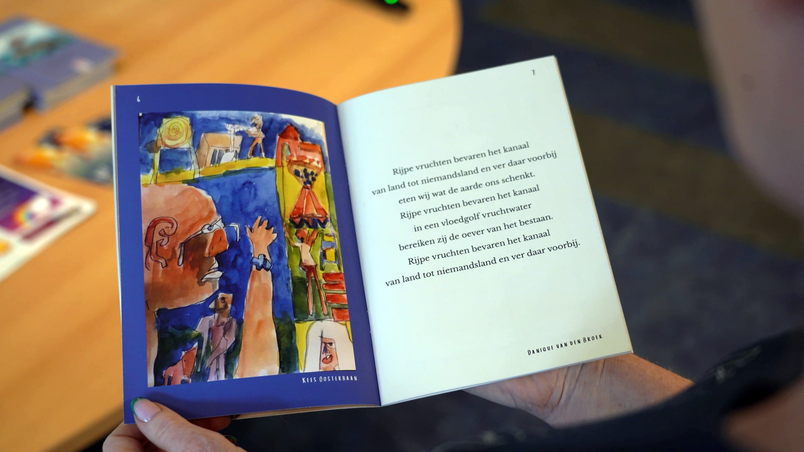 Handen houden een boek open met een illustratie aan de linkerkant en tekst aan de rechterkant. De illustratie toont kleurrijke abstracte figuren. De tekst begint met "Rijpe vruchten bevaren het kanaal" en is geschreven in het Nederlands. Het boek ligt op een houten tafel met andere boeken op de achtergrond.