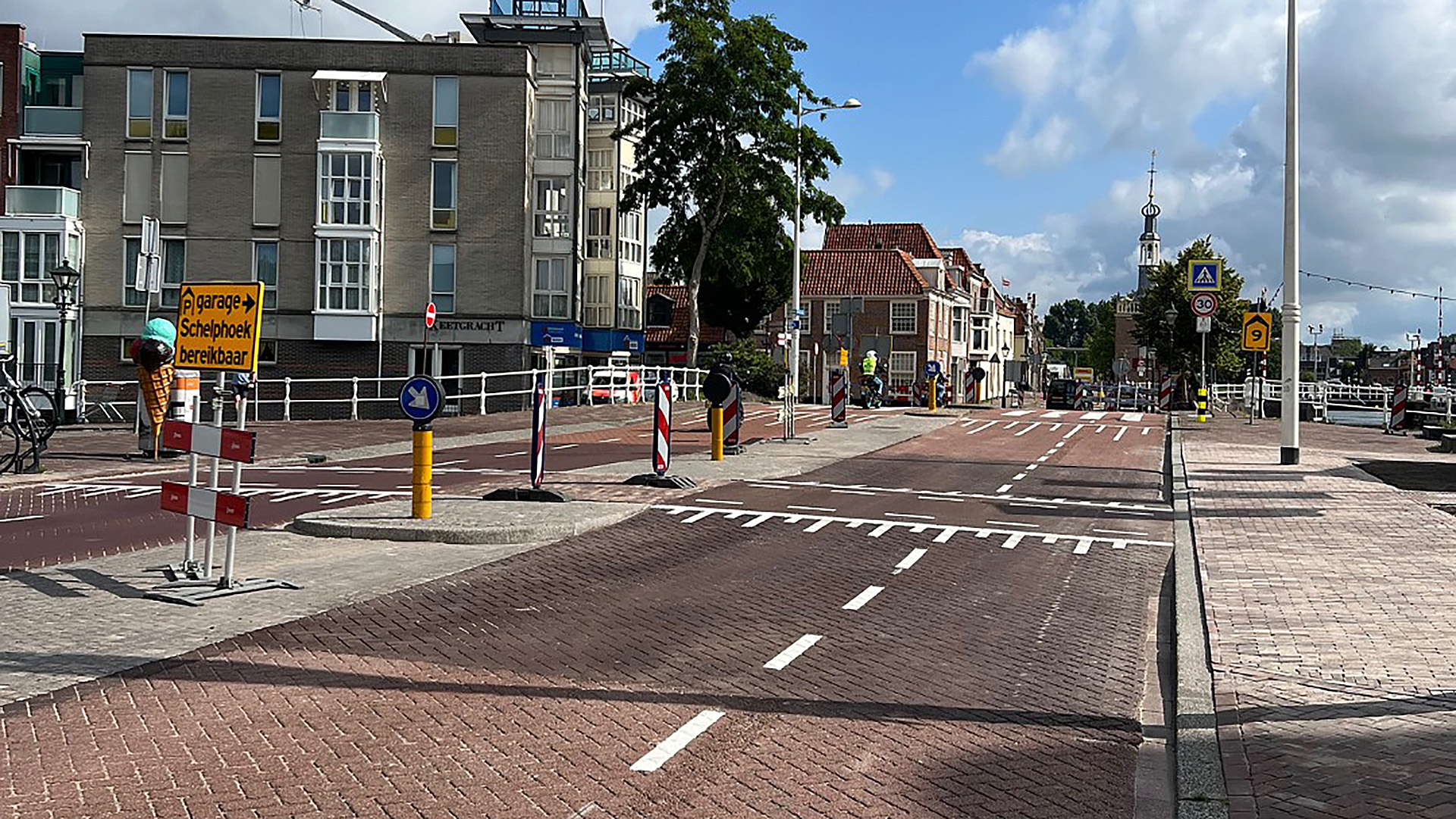 Weg met verkeersborden en markeringen in een stedelijke omgeving, met een wegwijzer naar parkeergarage Schelphoek, en enkele gebouwen en een kerktoren op de achtergrond.