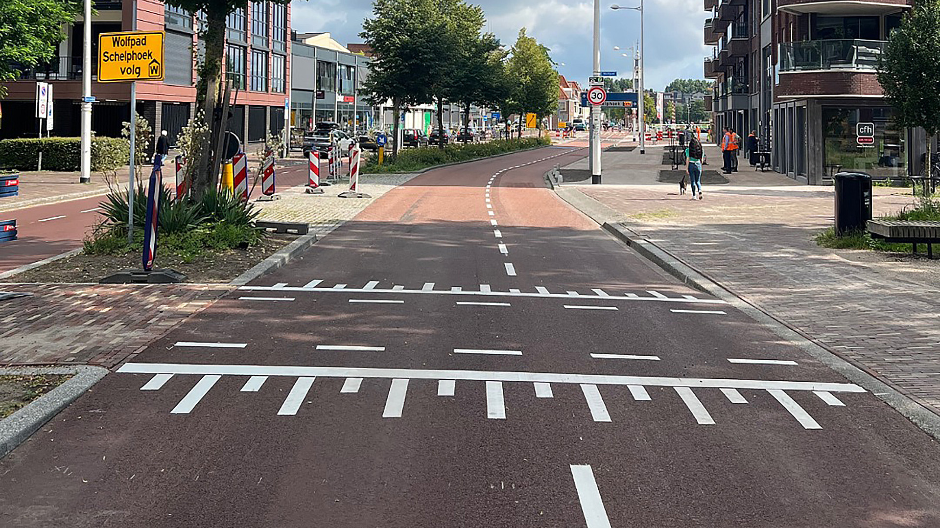 Straatbeeld met een gescheiden fietspad en bord met tekst 'Wolfpad Schelphoek volg', omringd door stedelijke bebouwing en verkeersborden.