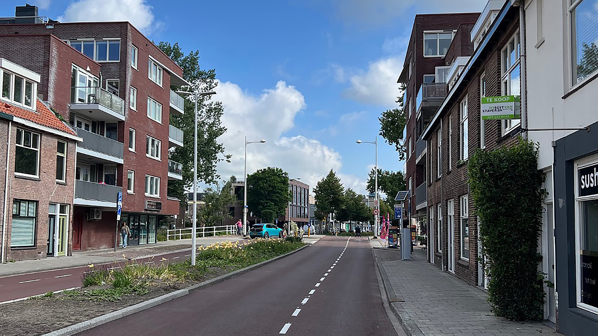 Straatbeeld met rijtjeshuizen en appartementen aan beide zijden, een smalle rijstrook in het midden, en een auto in de verte. De lucht is blauw met enkele wolken en er staan bomen langs de straat.