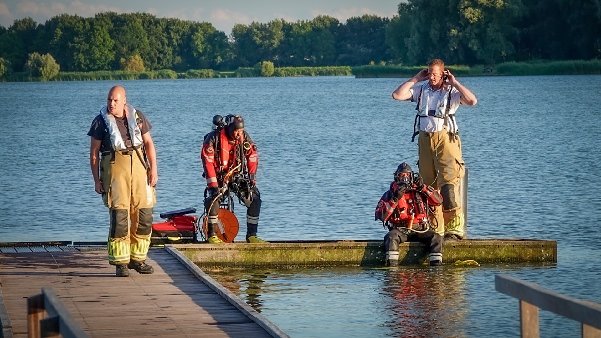 Brandweerlieden staan bij een meer; twee dragen duikuitrusting en zitten op een steiger, terwijl twee anderen staan en beschermende uitrusting dragen.