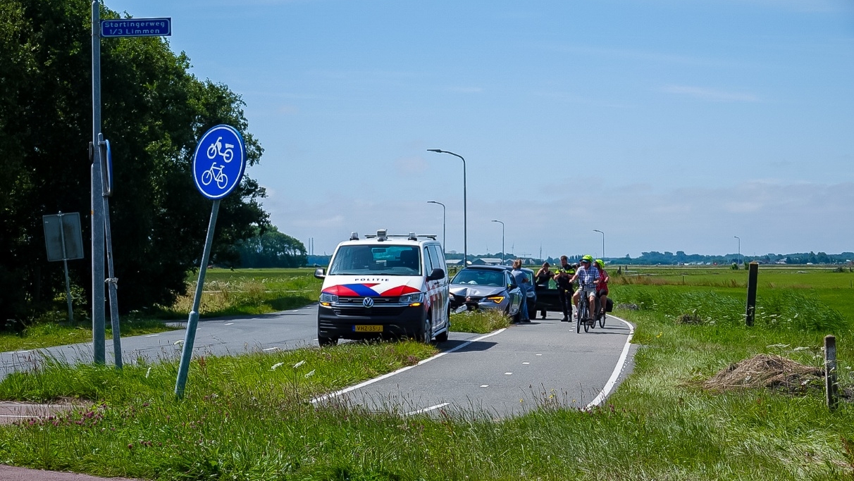 Een fietspad in een landelijke omgeving met een politieauto en een andere auto geparkeerd langs de weg. Er zijn mensen aanwezig, inclusief een paar fietsers die voorbij rijden, en een verkeersbord dat aangeeft dat het een fietspad is.