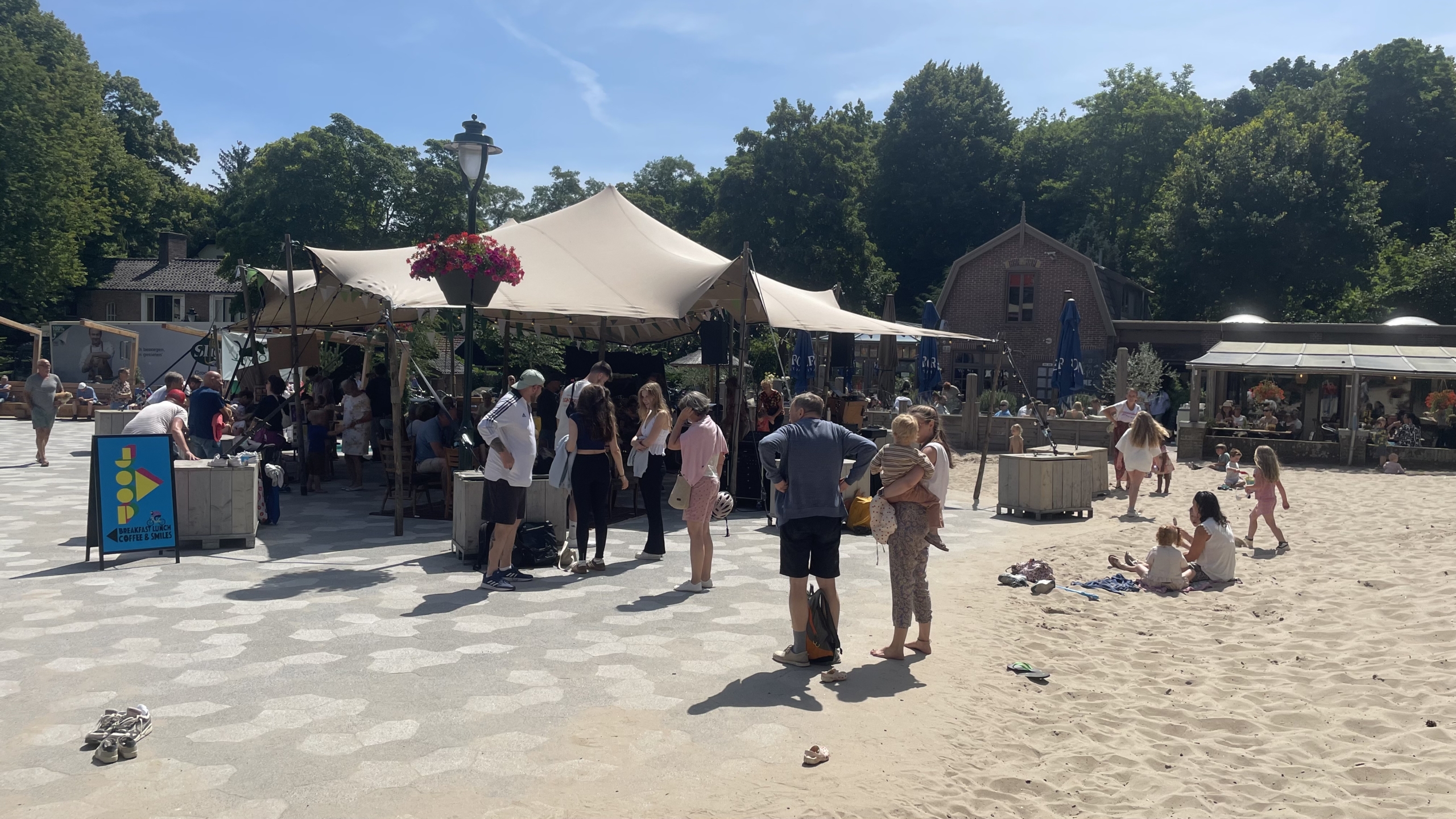 Mensen verzamelen zich bij een open tent met schaduw op een zonnige dag, met kinderen die in het zand spelen en een café op de achtergrond.