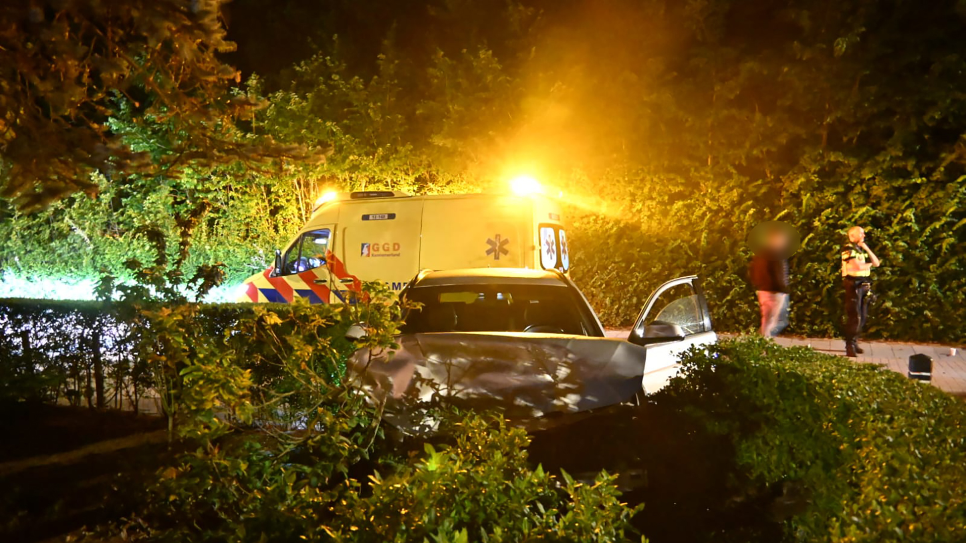 Een auto is gecrasht in de bosjes langs een weg, met een ambulance die met zwaailichten aanwezig is en een politieagent die met iemand praat op de achtergrond.