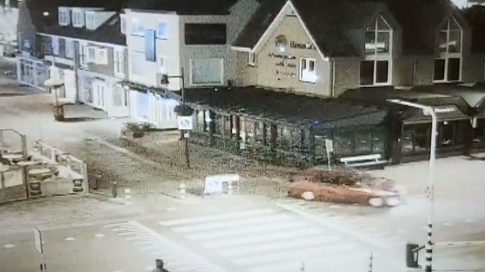 Een rood voertuig rijdt 's nachts over een kruising nabij een rij gebouwen met winkels en huizen.