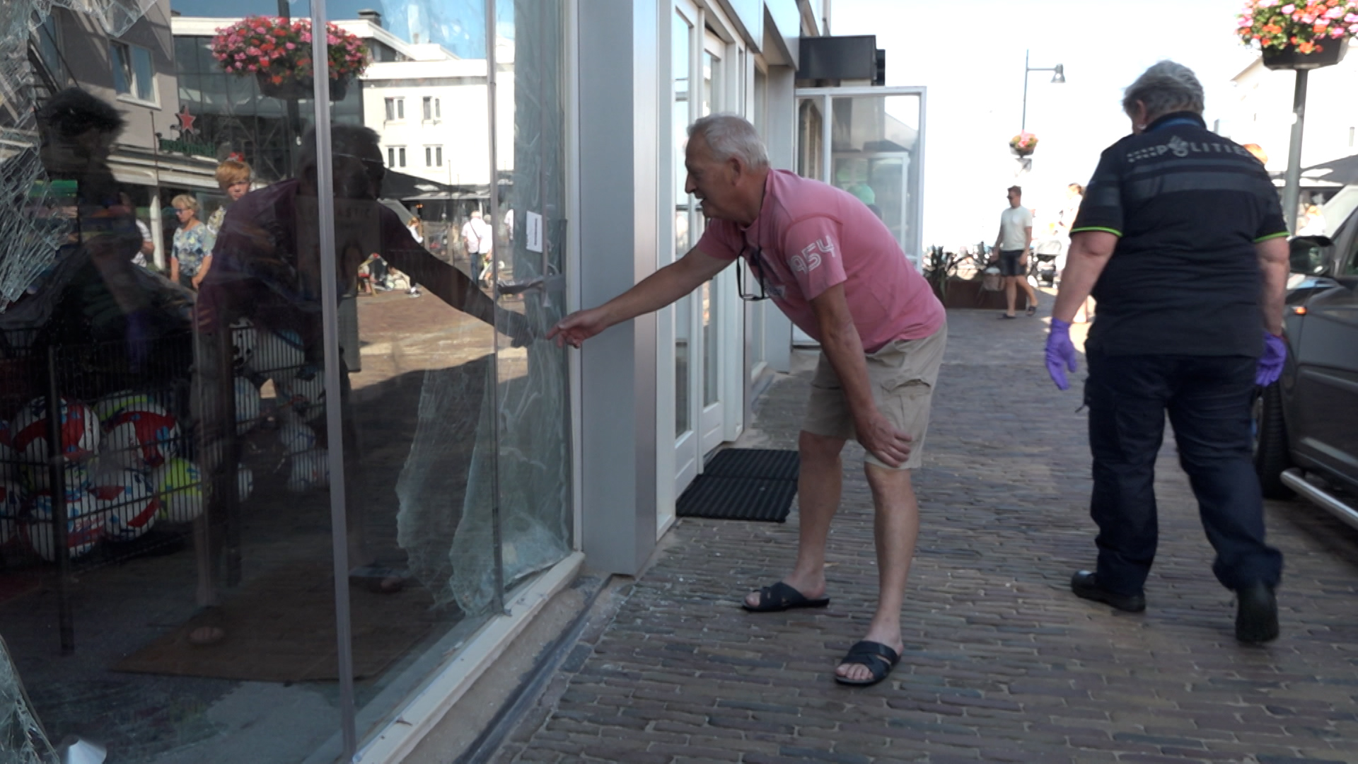 Een oudere man in een roze T-shirt inspecteert een gebroken glazen winkelruit, terwijl een persoon in uniform met paarse handschoenen erbij staat op een zonnige dag in een winkelstraat.