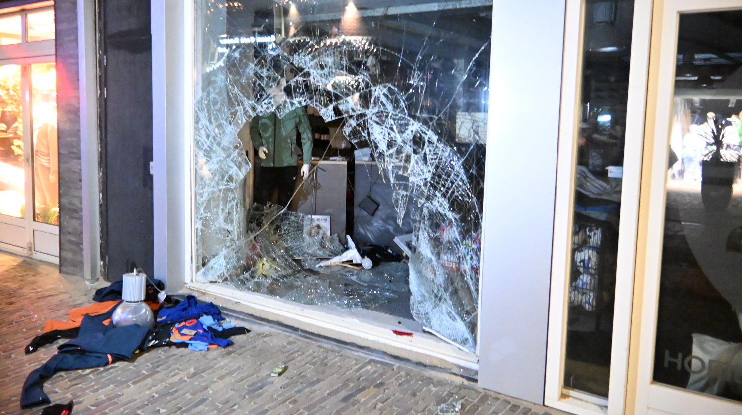 Gebroken winkelruit met een grote opening in het glas, omringd door scherven en kleding op de grond; binnen staat een mannequin. Het vindt plaats naast een winkel met accessoires, te zien vanaf de straat.