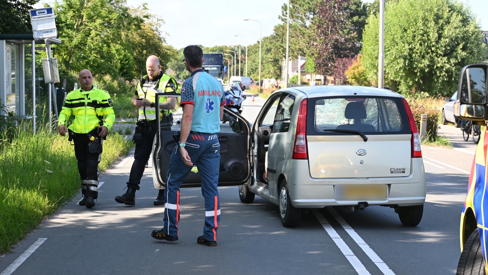 Twee politieagenten en een ambulancepersoneelslid staan naast een zilverkleurige auto op een weg; de agenten overleggen terwijl de ambulancepersoneelslid naar de open bestuurdersdeur kijkt.