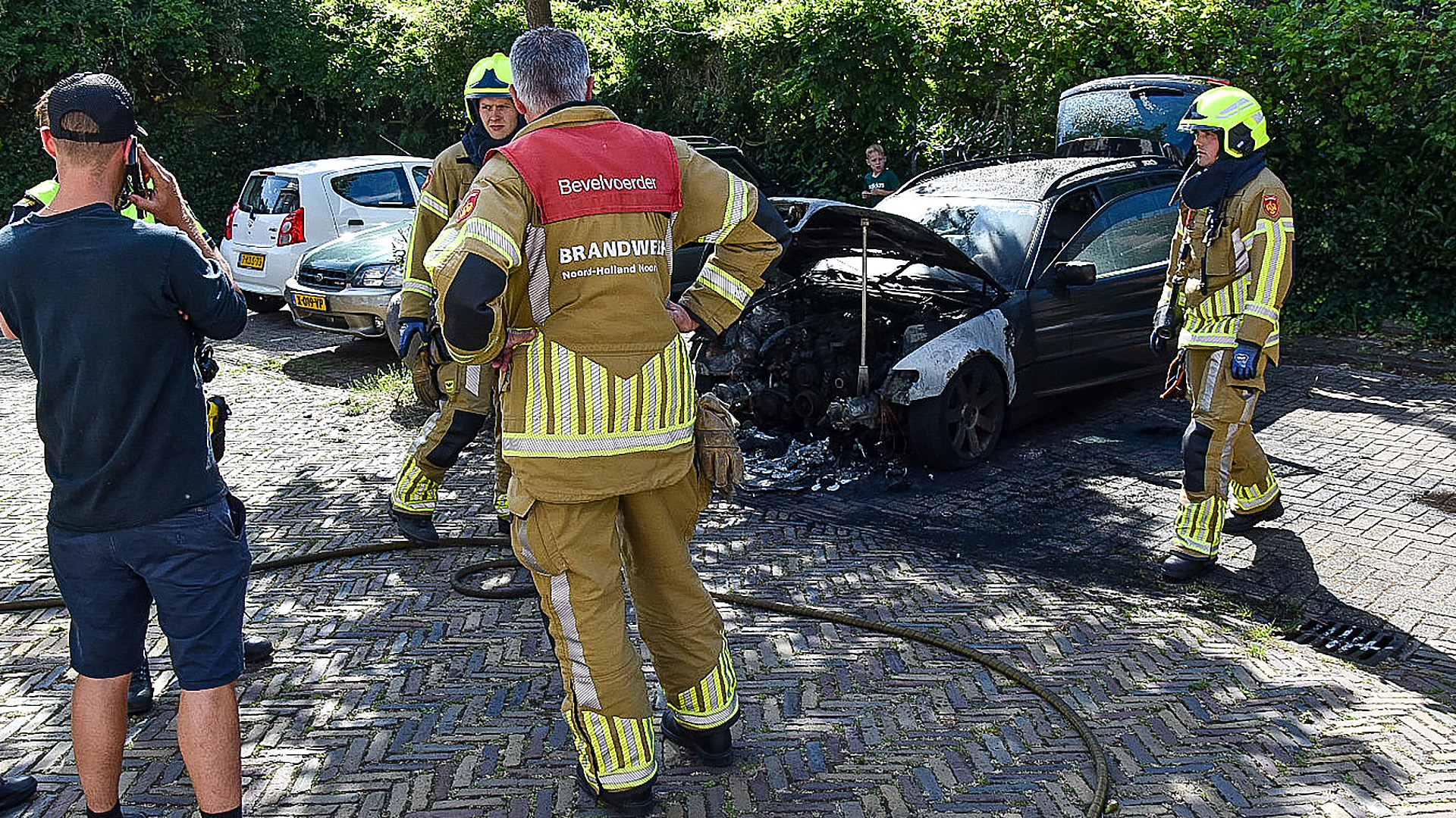 Brandweermannen in uniform onderzoeken een beschadigde auto met een verbrande motorkap op een klinkerweg, omgeven door andere geparkeerde auto's en groene struiken.