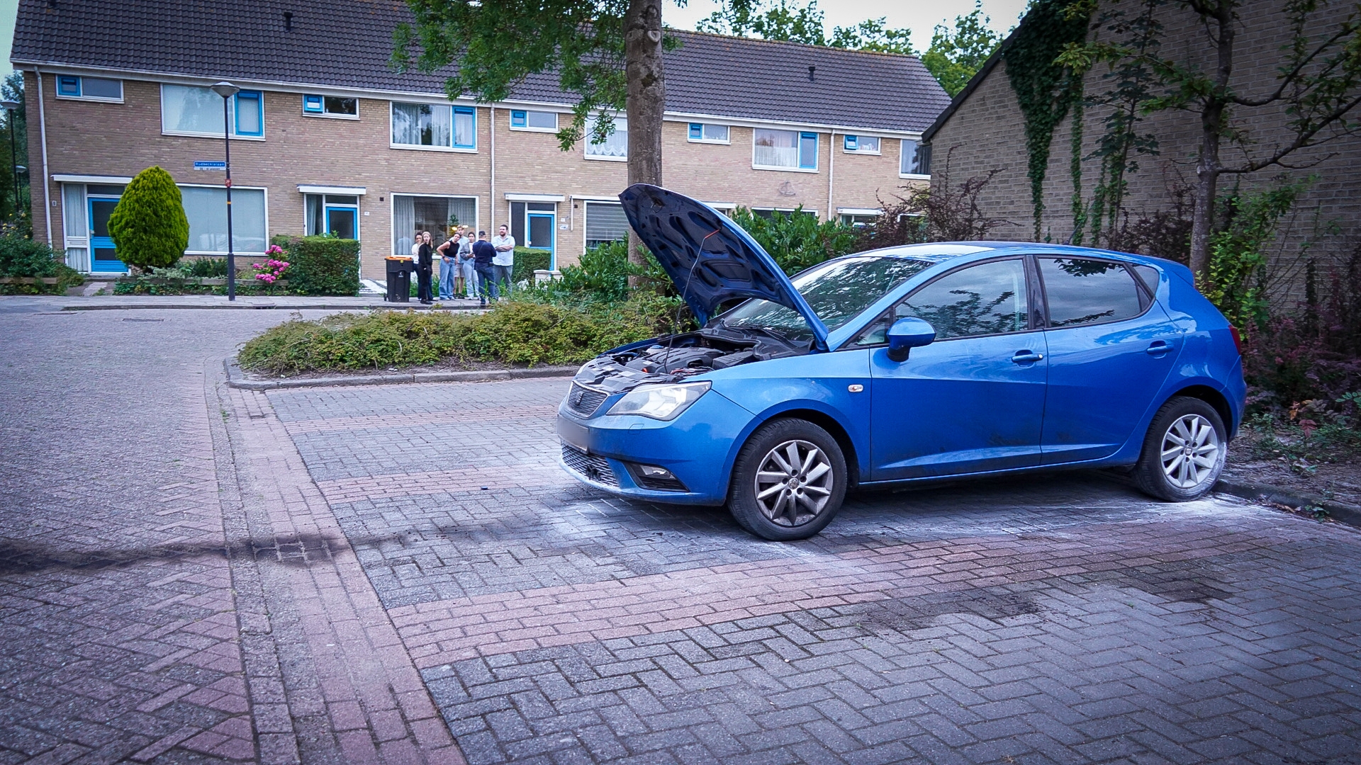 Een blauwe auto met geopende motorkap staat geparkeerd op een straat in een woonwijk, terwijl een groep mensen vanaf een afstand toekijkt.