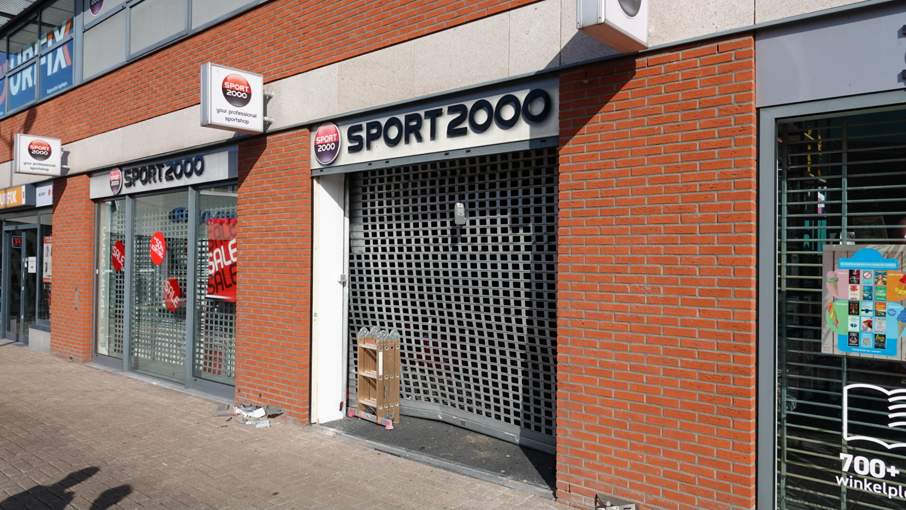 Een sportwinkel met de naam SPORT 2000, met een gesloten en beschadigd rolluik aan de voorzijde, en een rode baksteenmuur.