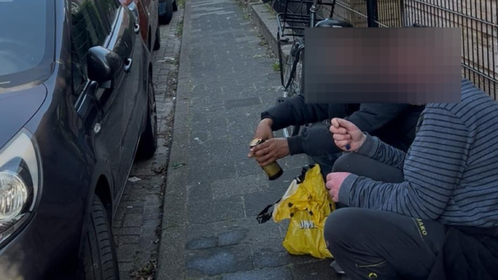 Twee mensen zitten op een stoep naast een zwarte auto; een van hen houdt een bierfles vast en er ligt een gele plastic tas naast hen.