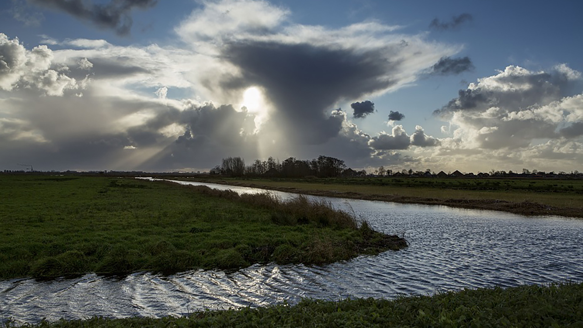 Landschap met een kronkelende rivier, groene grasvelden en een dramatische bewolkte hemel met zonnestralen die door de wolken breken.