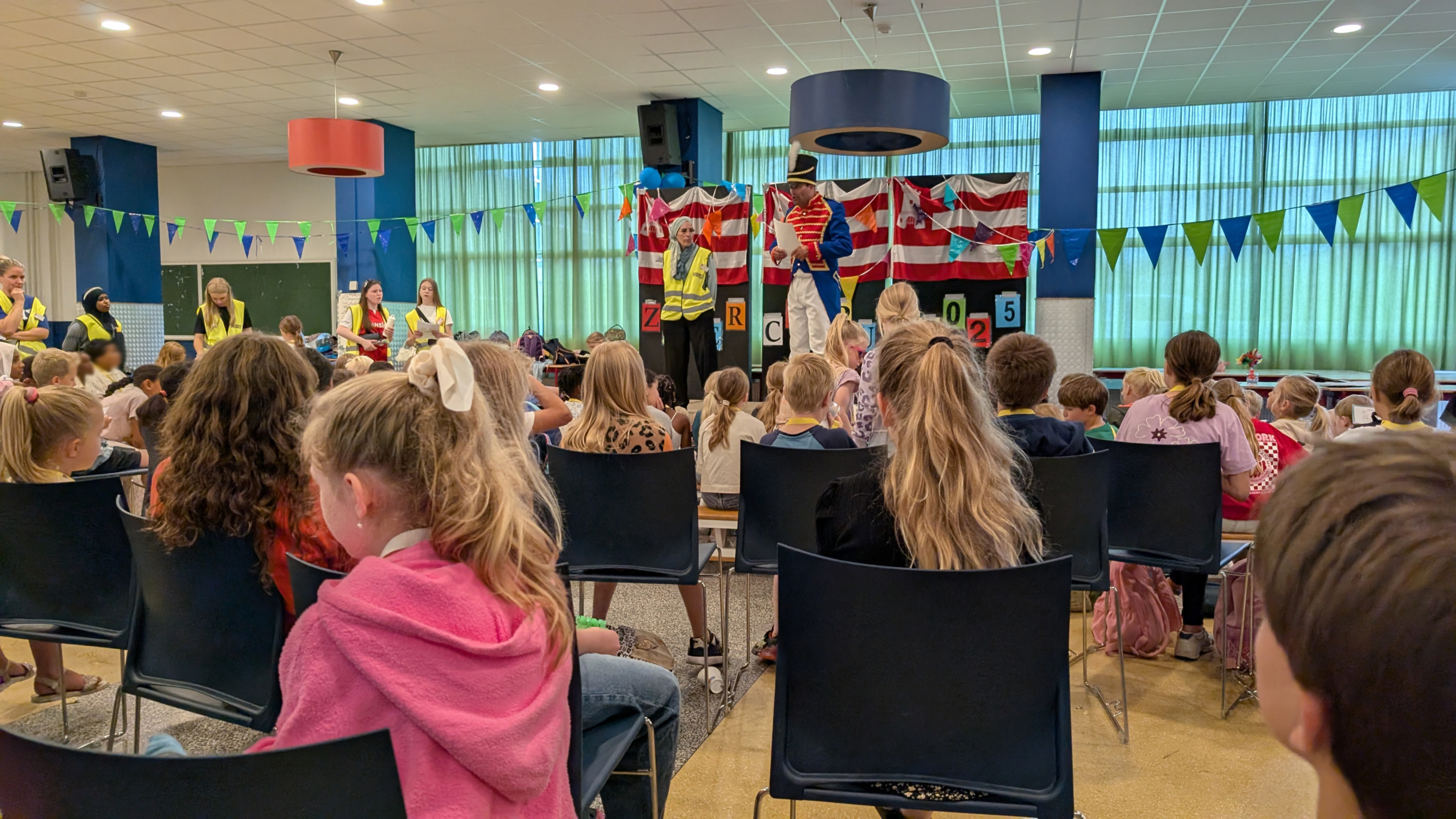 Kinderen zitten in stoelen en kijken naar een toneelstuk op een podium met kleurrijke decoraties en mensen in kostuums, waaronder iemand in een blauw uniform en een hoed. Er hangen vlaggetjes en enkele volwassenen dragen gele hesjes.