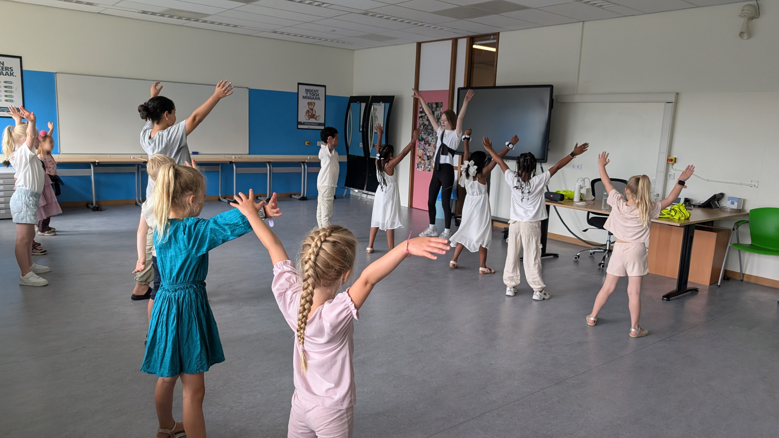 Een groep kinderen en een volwassene neemt deel aan een dansles in een klaslokaal, waarbij iedereen zijn armen in de lucht houdt.