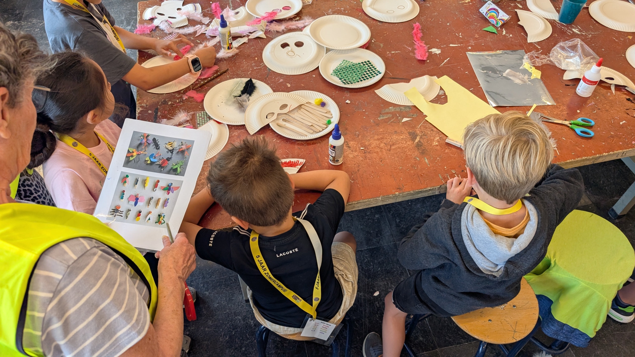 Kinderen zitten aan een tafel met knutselmaterialen zoals papieren bordjes, lijm, veertjes, en gekleurd papier, terwijl een volwassene een blad met knutselideeën vasthoudt.