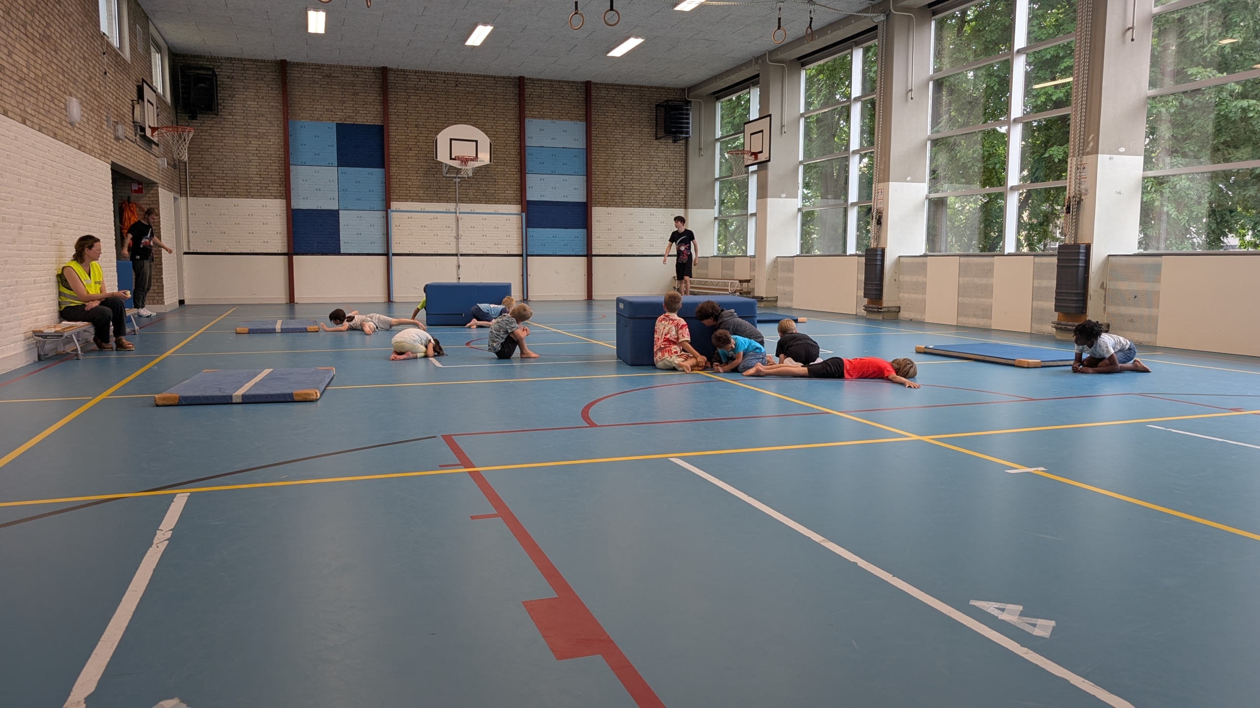 Kinderen spelen in een gymzaal, liggend op de grond of op matten, terwijl enkele volwassenen toekijken.