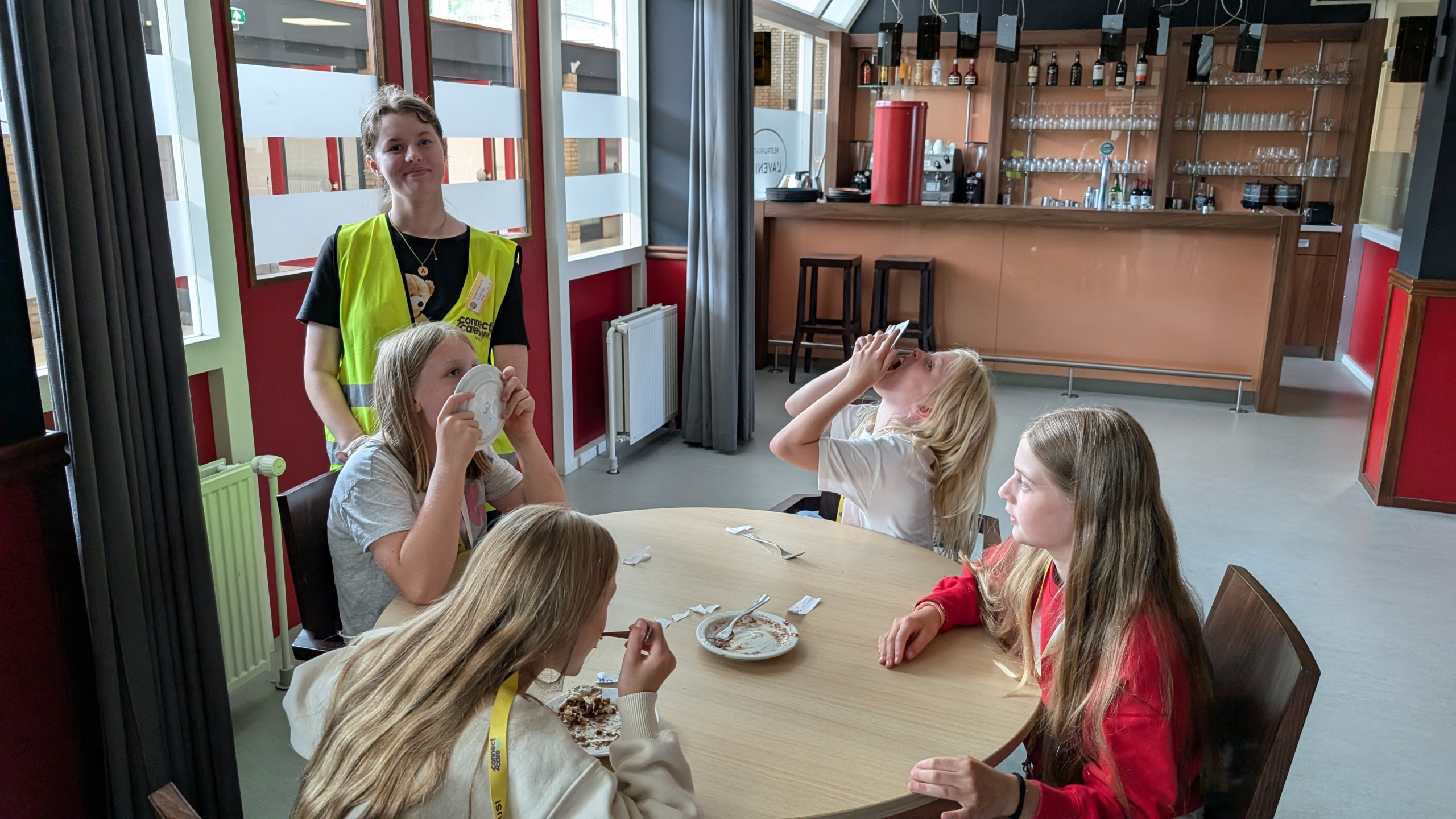 Vier kinderen zitten aan een tafel in een café-omgeving, met een volwassen persoon bij hen staande, gekleed in een geel veiligheidsvest. Drie kinderen eten van bordjes, waarvan twee borden vasthouden aan hun mond, terwijl de derde een lepel gebruikt. De vierde persoon kijkt recht vooruit.