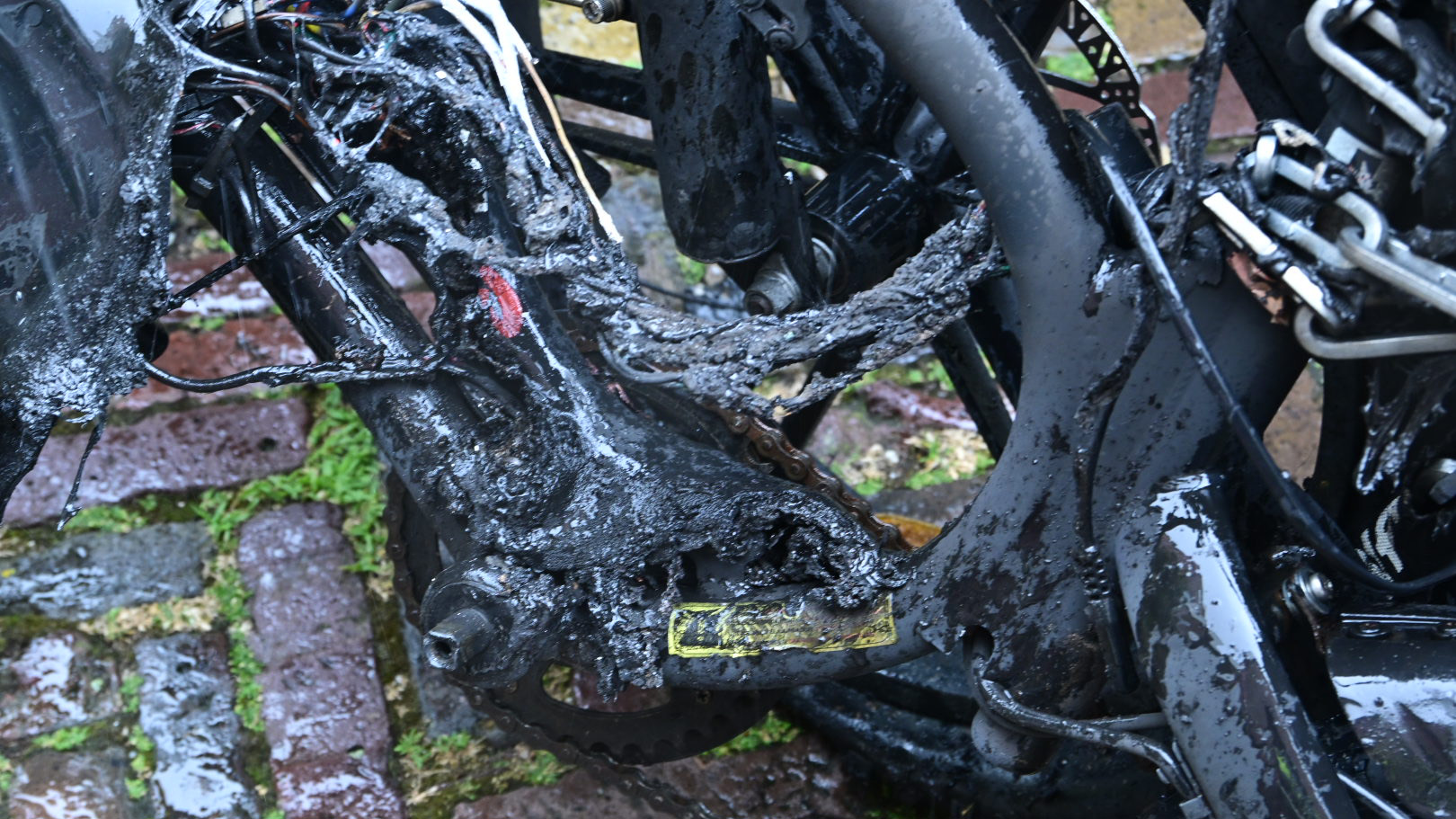 Een close-up van een zwart frame van een fiets dat zwartgeblakerd en beschadigd is, met gesmolten en gescheurde kabels langs het frame en groene mos of gras tussen bakstenen ondergrond eronder.
