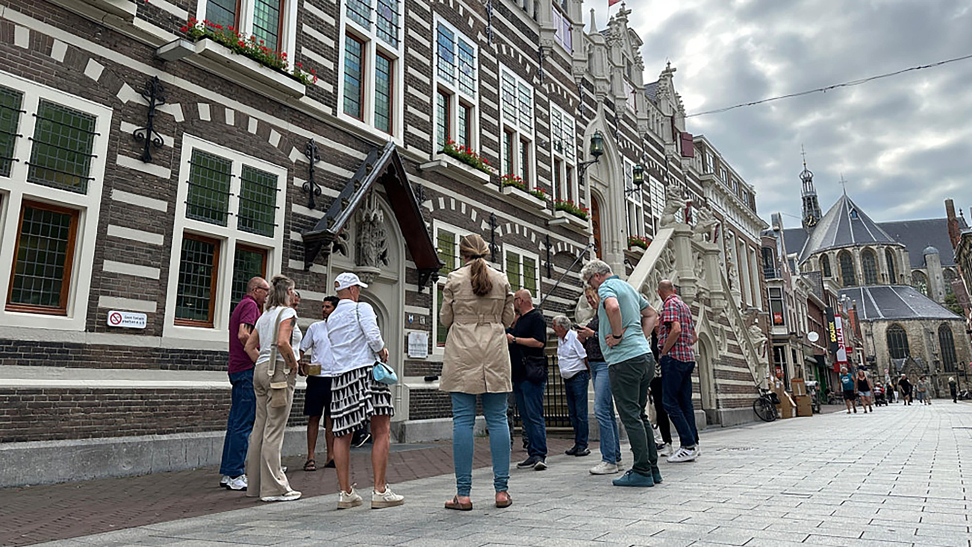 Een groep mensen verzameld voor een historisch gebouw met een gevel van baksteen en witte accenten, naast een kerk met een hoge torenspits op de achtergrond.