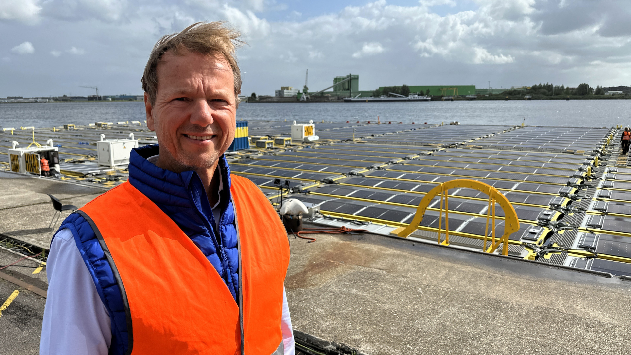 Man in an orange safety vest standing on a dock near an array of floating solar panels on the water, with an industrial area in the background.