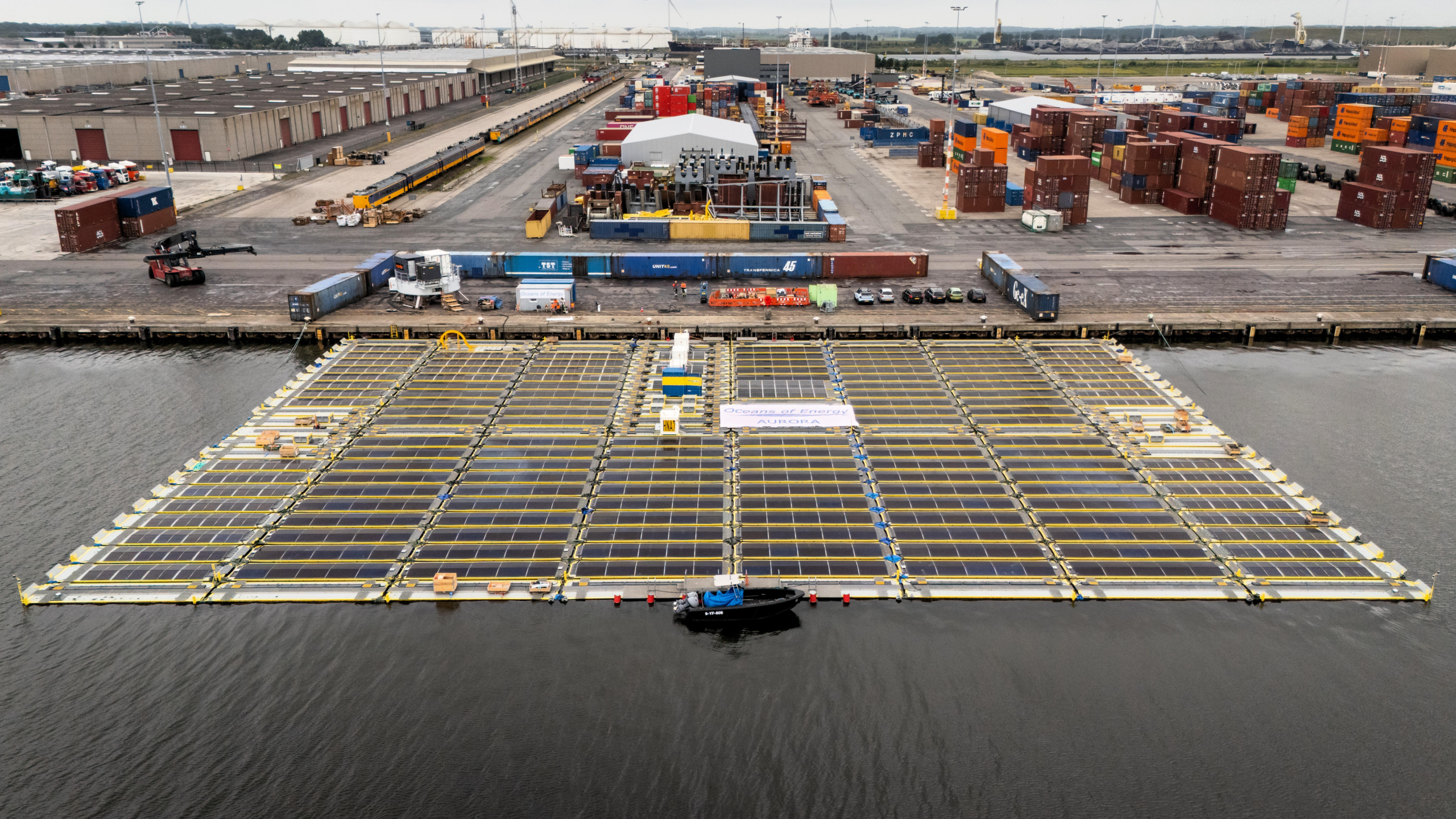 Zicht op een grote installatie van drijvende zonnepanelen in een havengebied, met stapels containers en opslaggebouwen op de achtergrond.