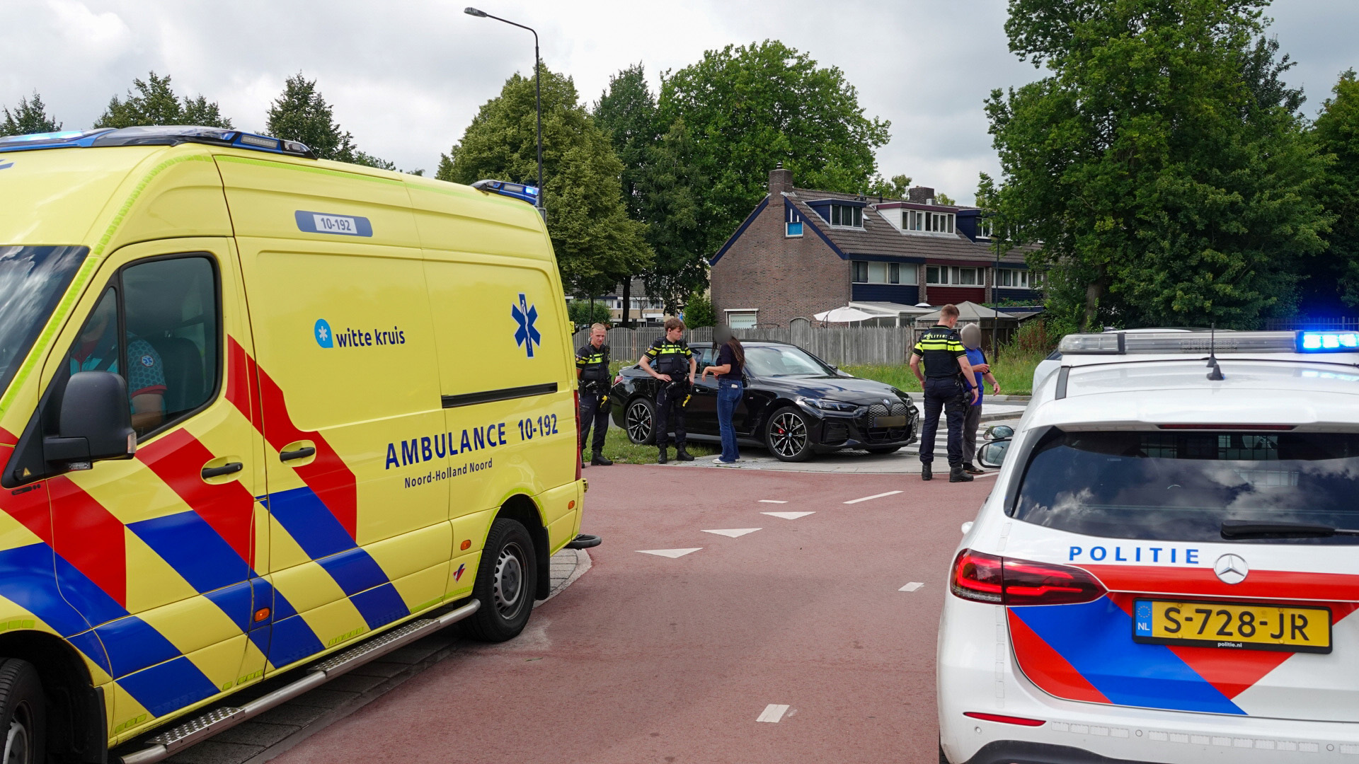 Een gele ambulance en een politieauto staan geparkeerd op de weg in een woonwijk, met agenten en een vrouw naast een zwarte auto. Op de achtergrond zijn bomen en een huis zichtbaar.