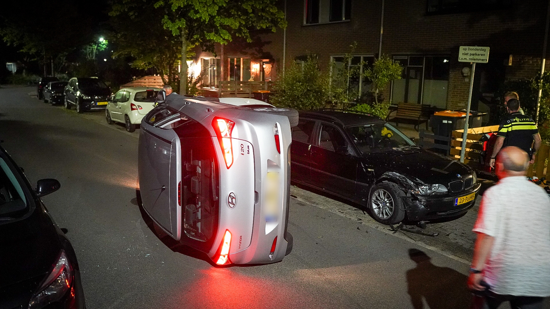 Auto ligt op zijn zijkant met de lichten aan in een woonstraat, naast andere geparkeerde auto's, met een politieagent aanwezig op de achtergrond.