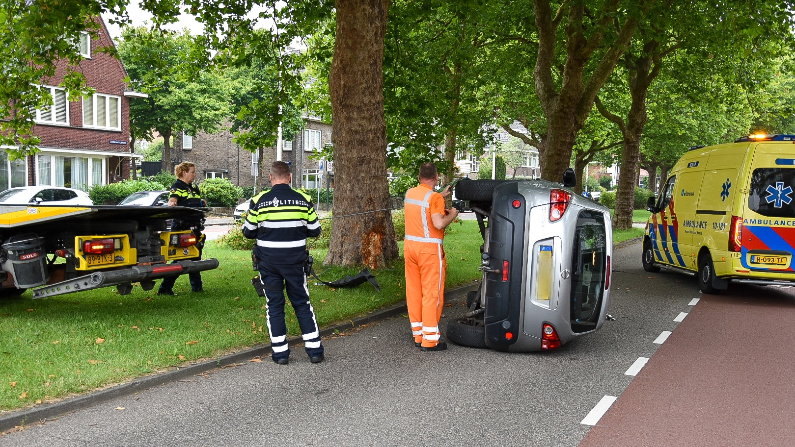 Een auto ligt op zijn zijkant op de weg naast een omgevallen boom, met politieagenten en een wegenwerker erbij. Er is een bergingswagen en een ambulance aanwezig. De omgeving is een straat met huizen en bomen.