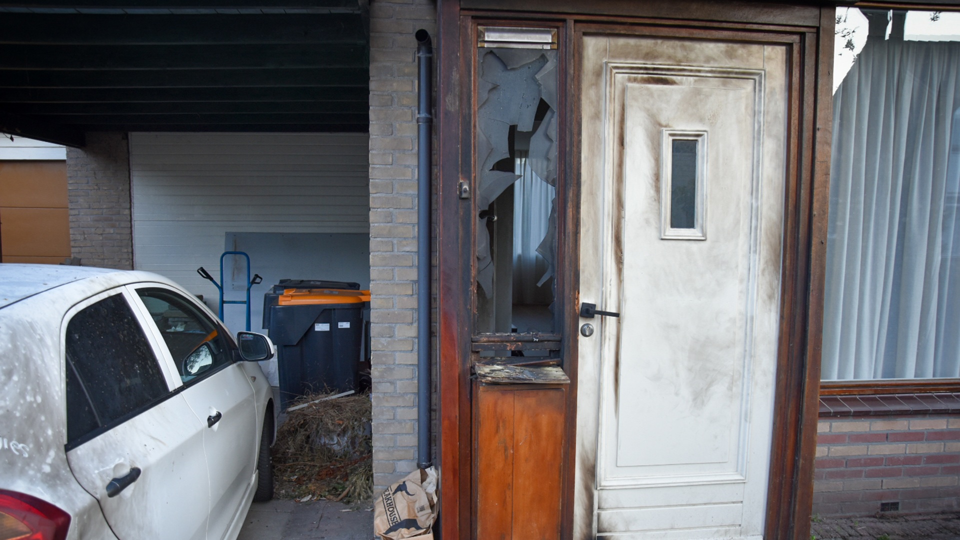 Een beschadigd gebouw met een kapot raam en een smoezelige deur, een witte auto geparkeerd ernaast, en vuilnisbakken op de achtergrond.
