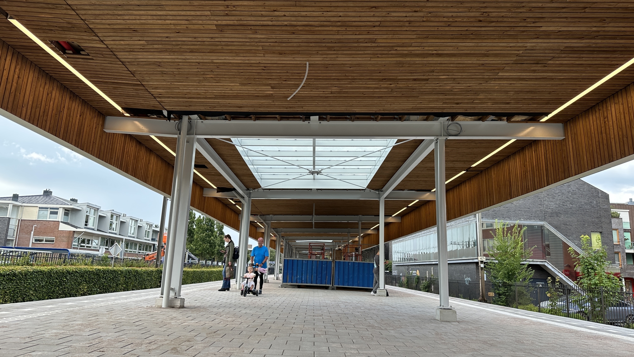 Overdekte wandelpassage met houten plafond en metalen constructie, waar enkele mensen lopen; aan de rechterkant staan een glazen gebouw en groene struiken.