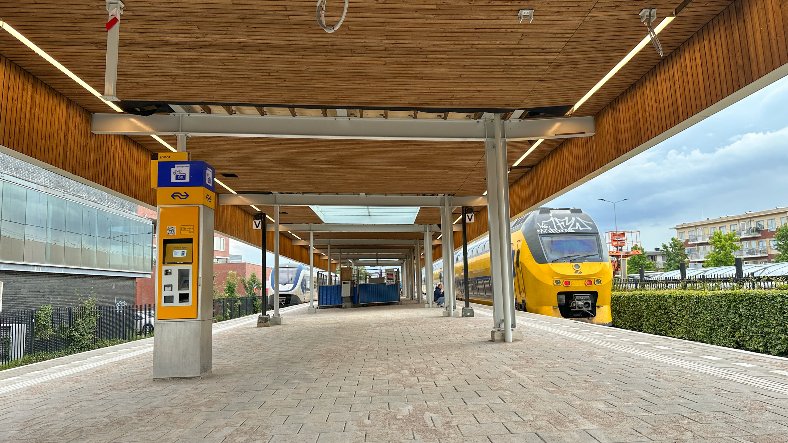 Een overdekt spoorwegperron in Nederland met een gele elektrische trein en een NS-kaartautomaat. Het houten plafond en de open omgeving zijn zichtbaar, met een wachtruimte in het midden van het perron.