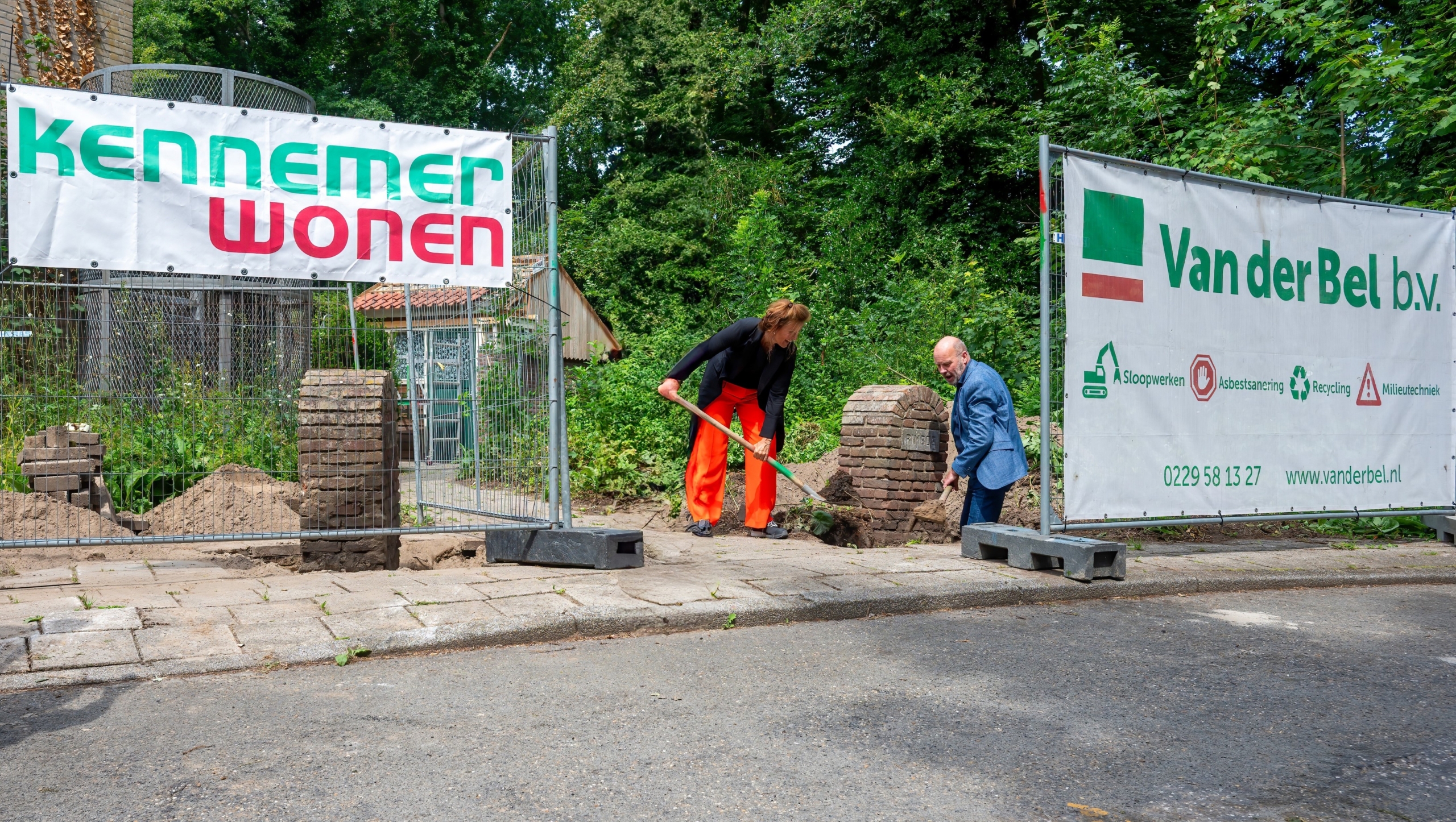 Twee mensen graven met scheppen naast een hek met spandoeken van Kennemer Wonen en Van der Bel b.v., op een trottoir omgeven door groen.