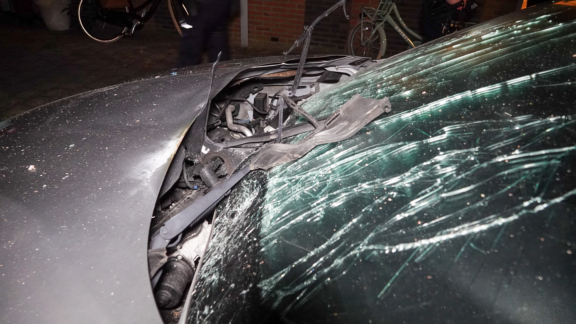 Een ernstig beschadigde auto met een ingedrukt motorkap en een gebarsten voorruit, omgeven door donkere nachtelijke verlichting.