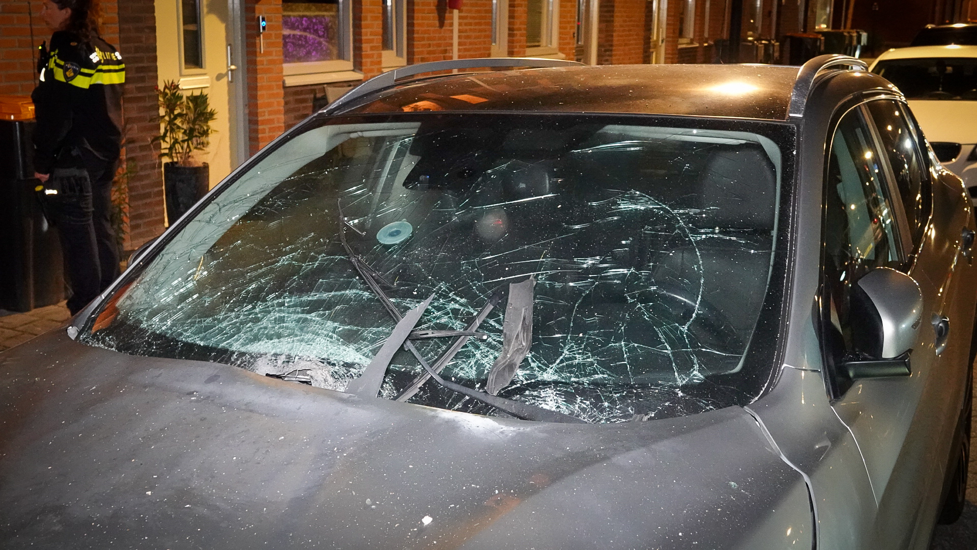 Auto met een zwaar beschadigde voorruit en ingedeukte motorkap, geparkeerd op straat naast een woning; een politieagent staat op de achtergrond.