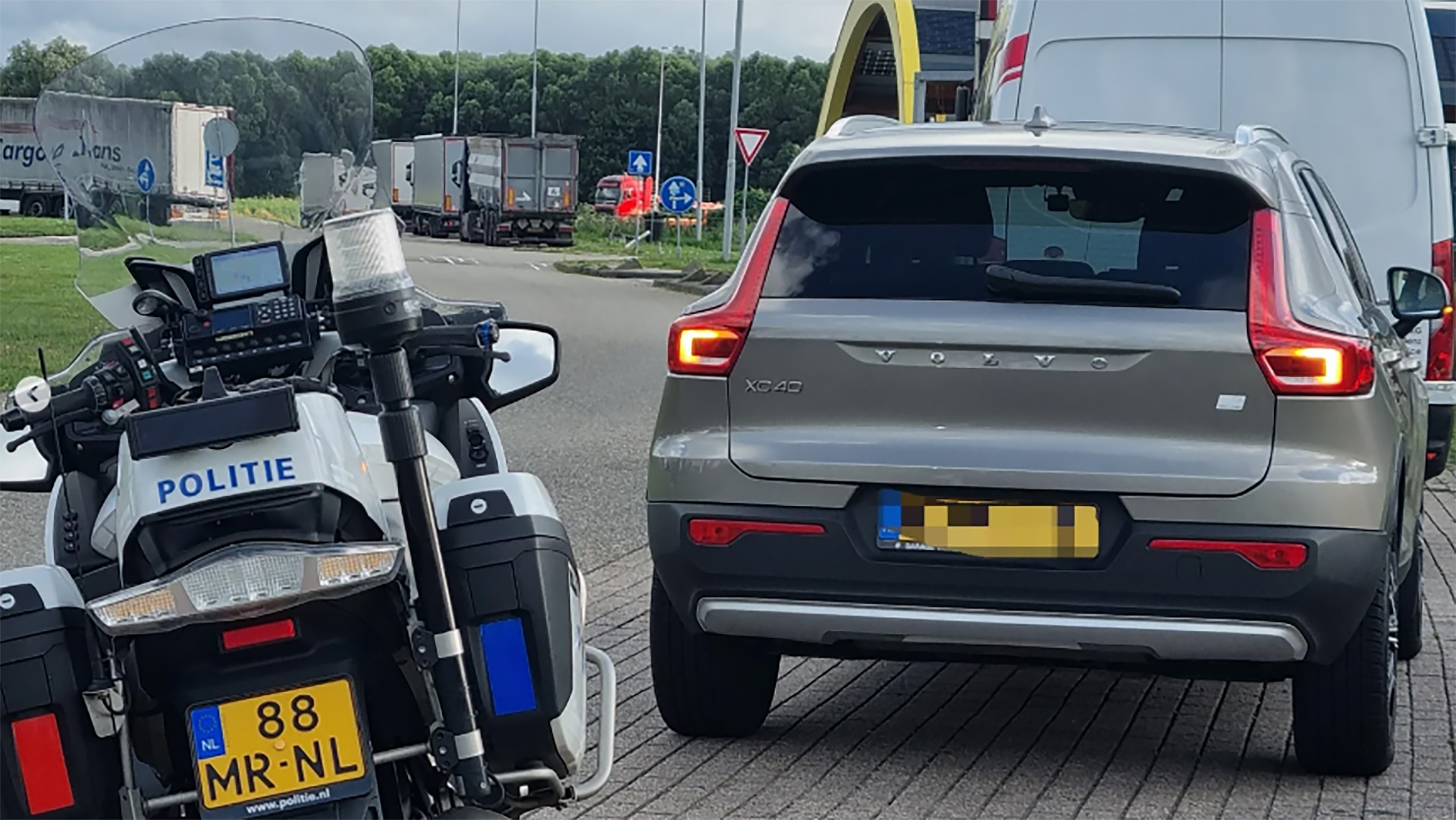 Een politie-motorfiets naast een geparkeerde zilverkleurige Volvo XC40 op een parkeerplaats met vrachtwagens op de achtergrond.