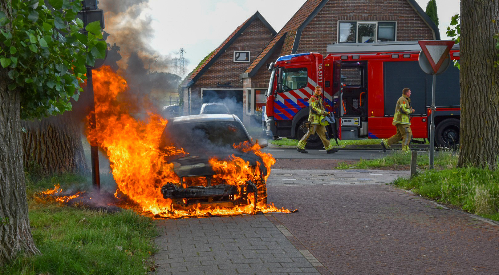 Een auto staat in brand op een stoep, omringd door felle vlammen. Op de achtergrond is een brandweerwagen te zien met brandweerlieden die zich naar de brandhaard haasten bij een woonhuis.