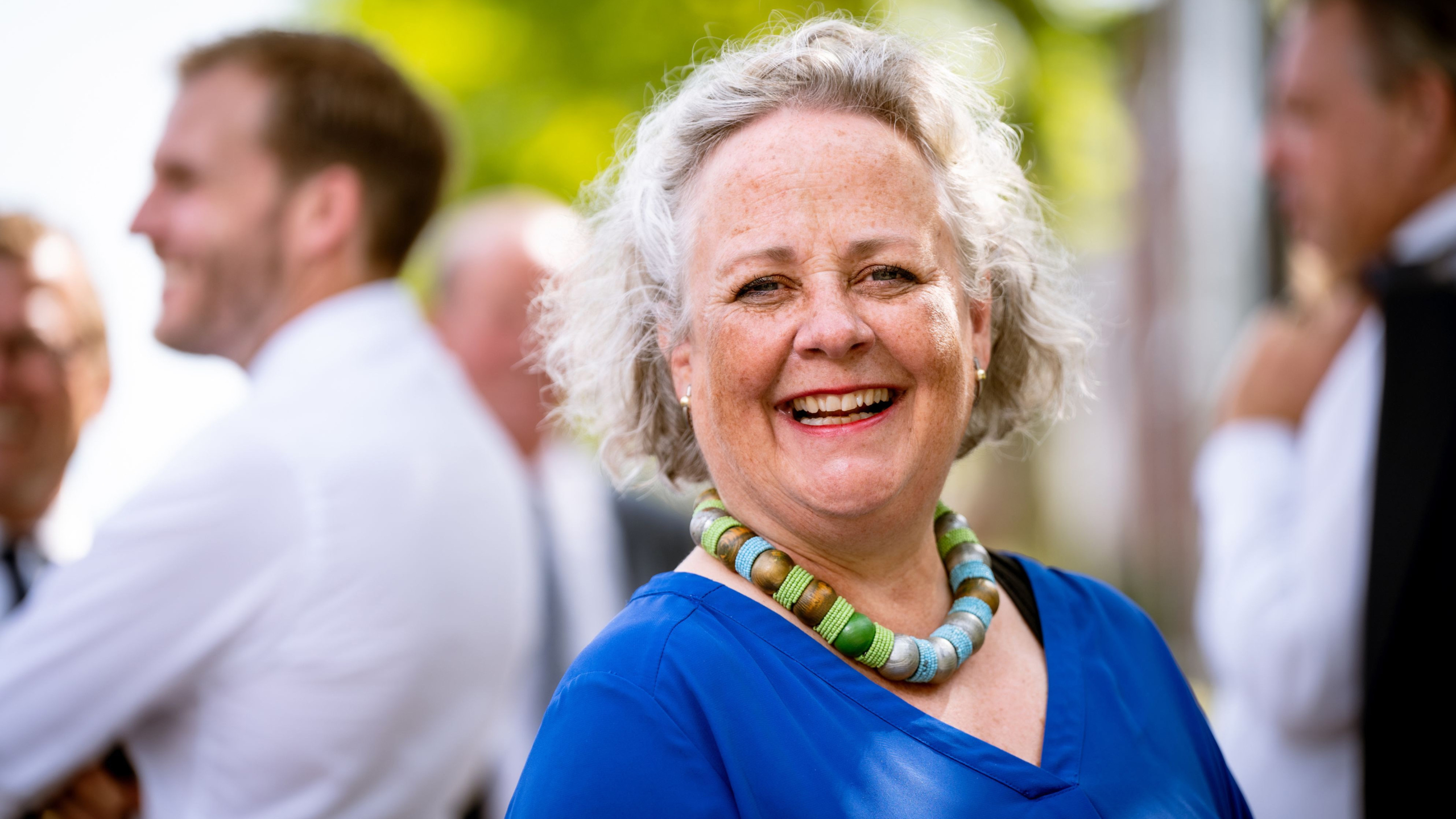 Lachende vrouw met grijs haar en een kleurrijke ketting, gekleed in een blauwe blouse, met mensen op de achtergrond op een zonnige dag.