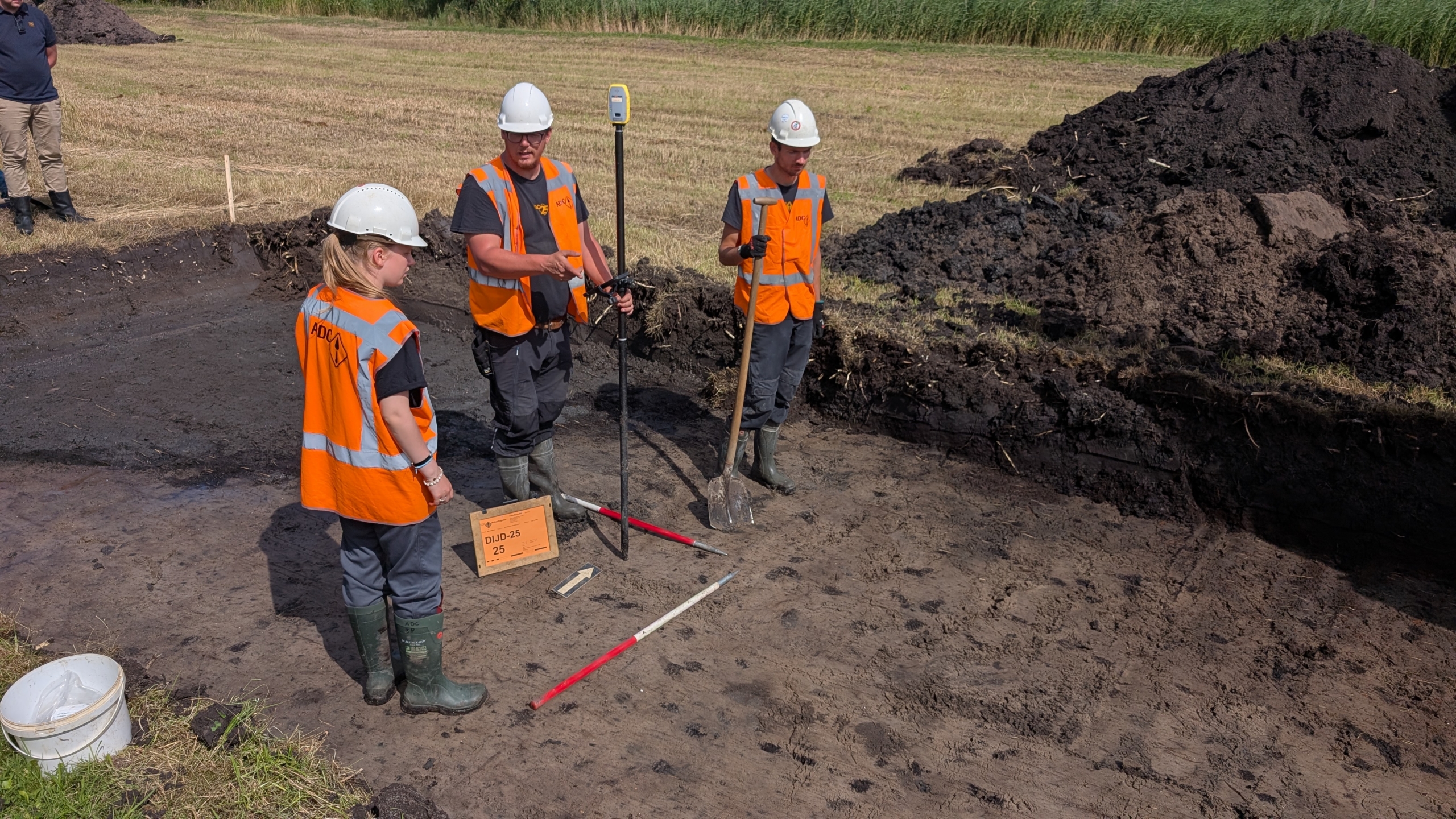 Drie archeologen in oranje veiligheidsvesten en witte helmen werken op een opgravingslocatie in een grasveld. Een man houdt een GPS-apparaat vast, terwijl een andere man een schep vasthoudt. De vrouw kijkt naar een bord met de tekst 'DIJD-25'. Er liggen hopen aarde om hen heen.