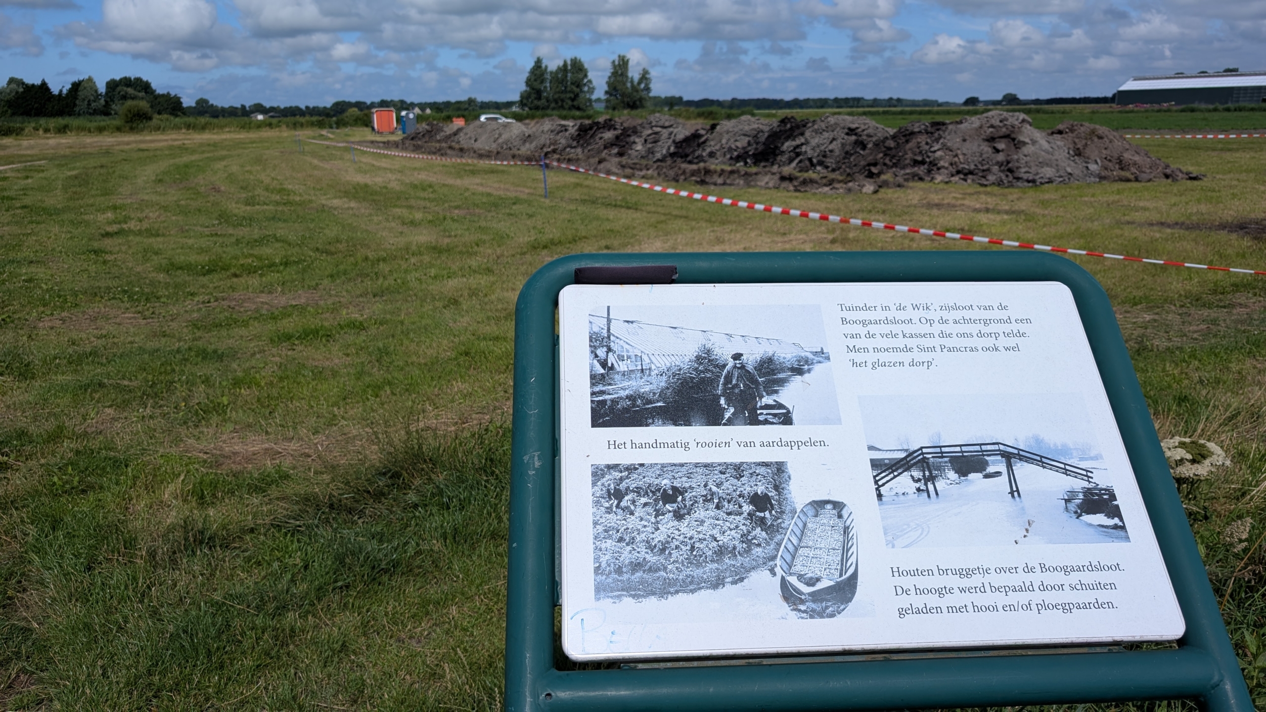 Informatiebord in een grasveld met foto's en tekst over de geschiedenis van het tuinieren in Sint Pancras, Nederland. Op de achtergrond zijn bouwvakkers en een stapel aarde te zien, afgezet met rood-wit lint.