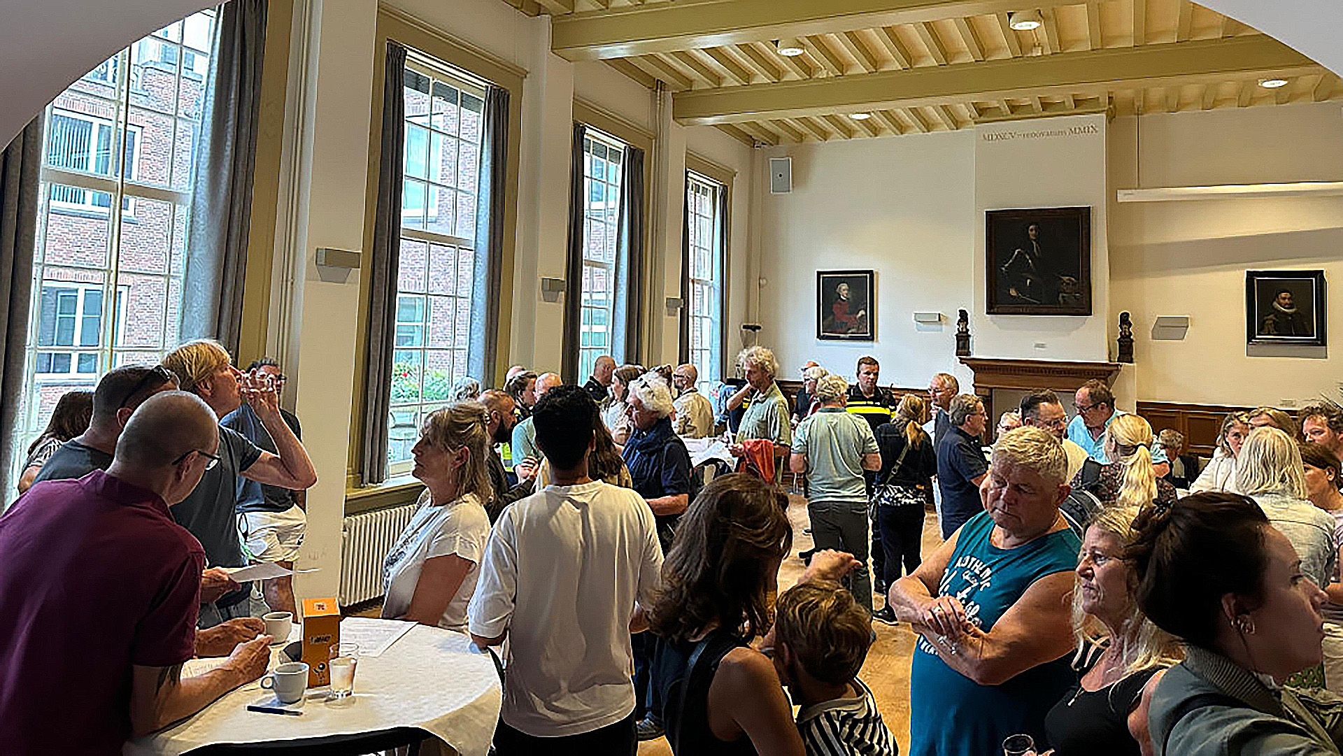 Een groep mensen verzameld in een kamer met hoge ramen en schilderijen aan de muren, bezig met sociale interacties en drinken.