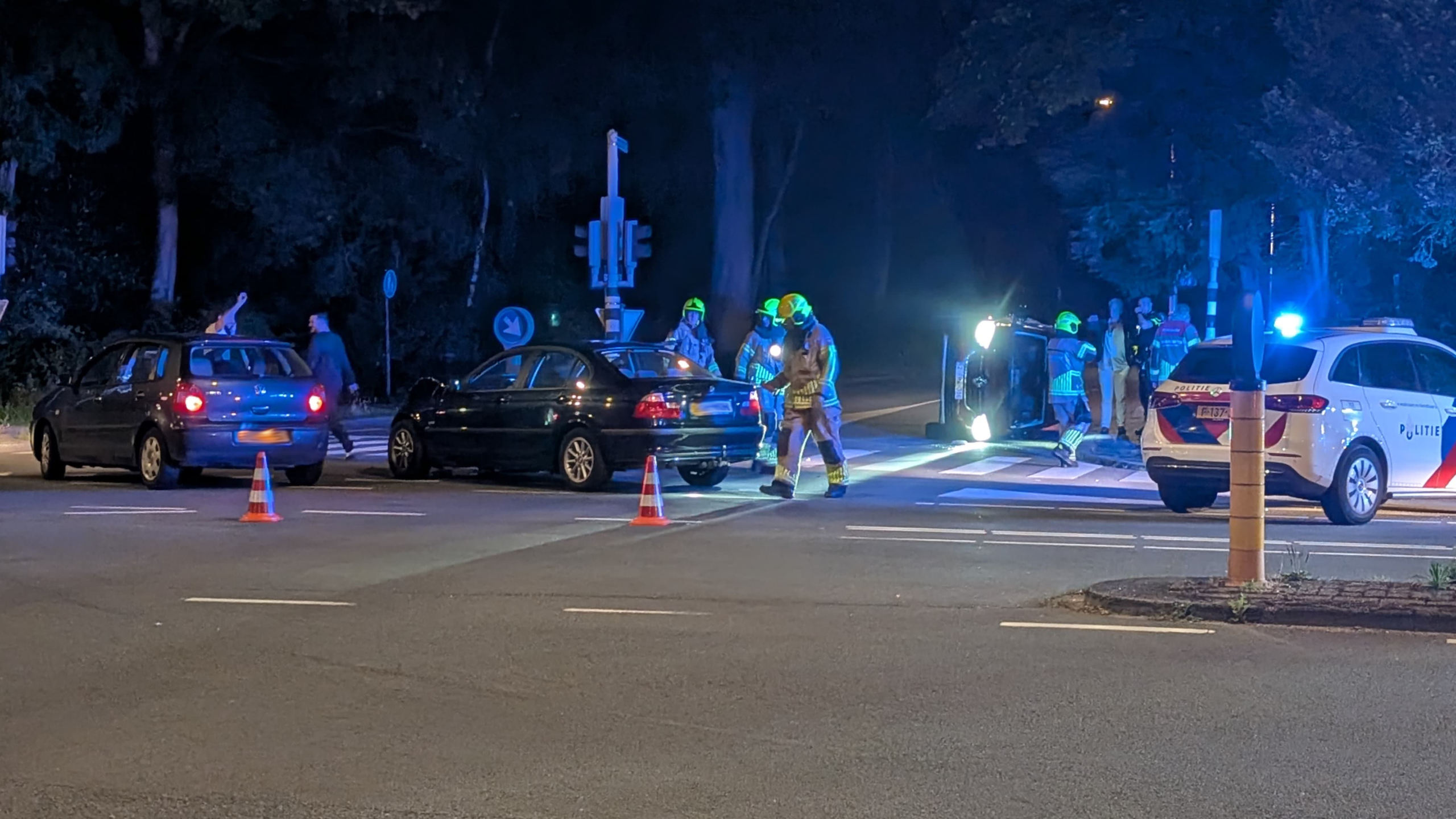 Een verkeersongeval met een omgevallen voertuig op een kruispunt, omringd door hulpverleners in reflecterende kleding. Er staan twee andere auto's en een politieauto met zwaailichten aan. Kegels markeren het gebied.