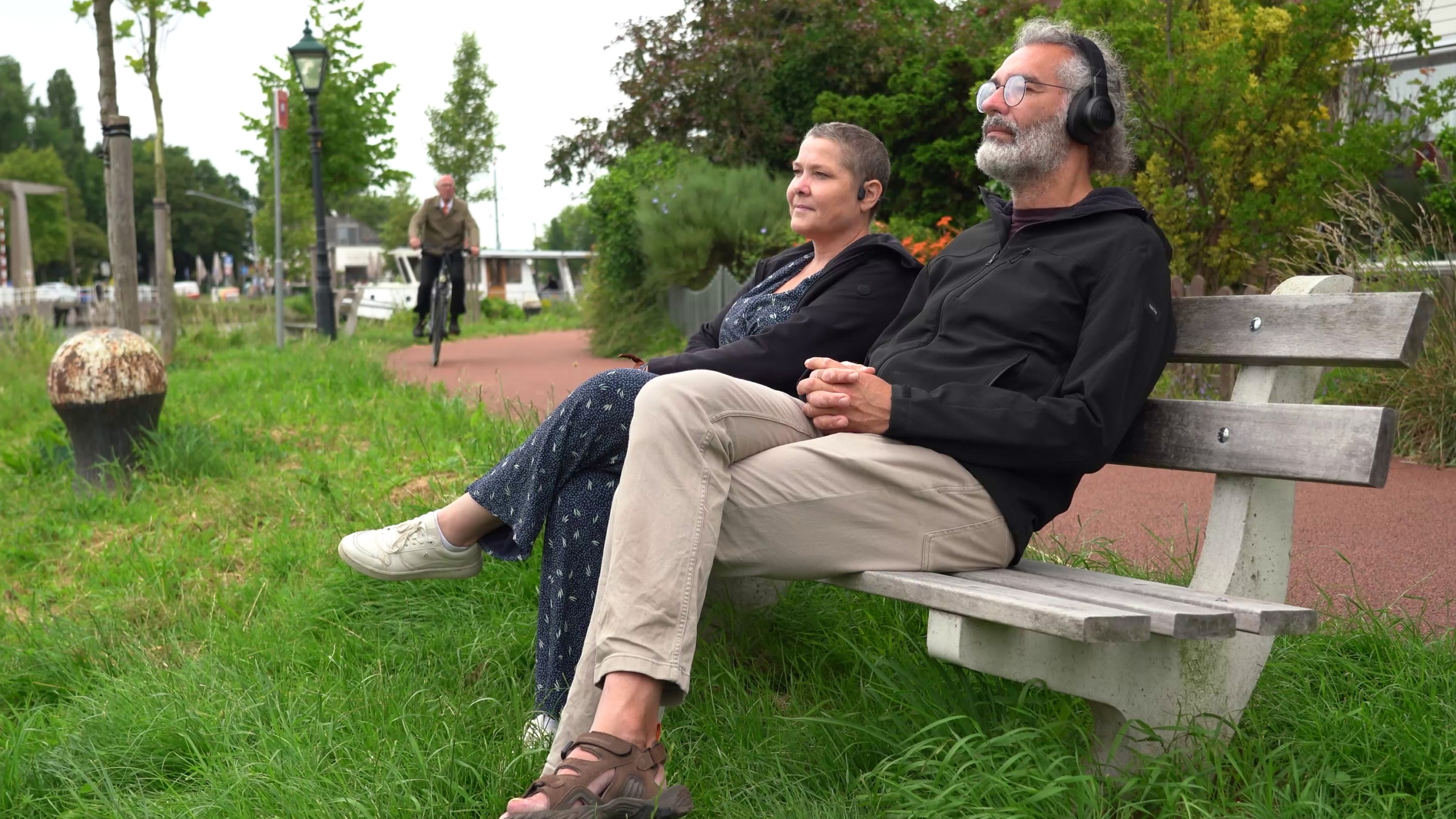 Twee mensen zitten ontspannen op een houten bankje langs een grasveld naast een fietspad, de man draagt een koptelefoon en beiden kijken voor zich uit, terwijl een fietser op de achtergrond passeert.
