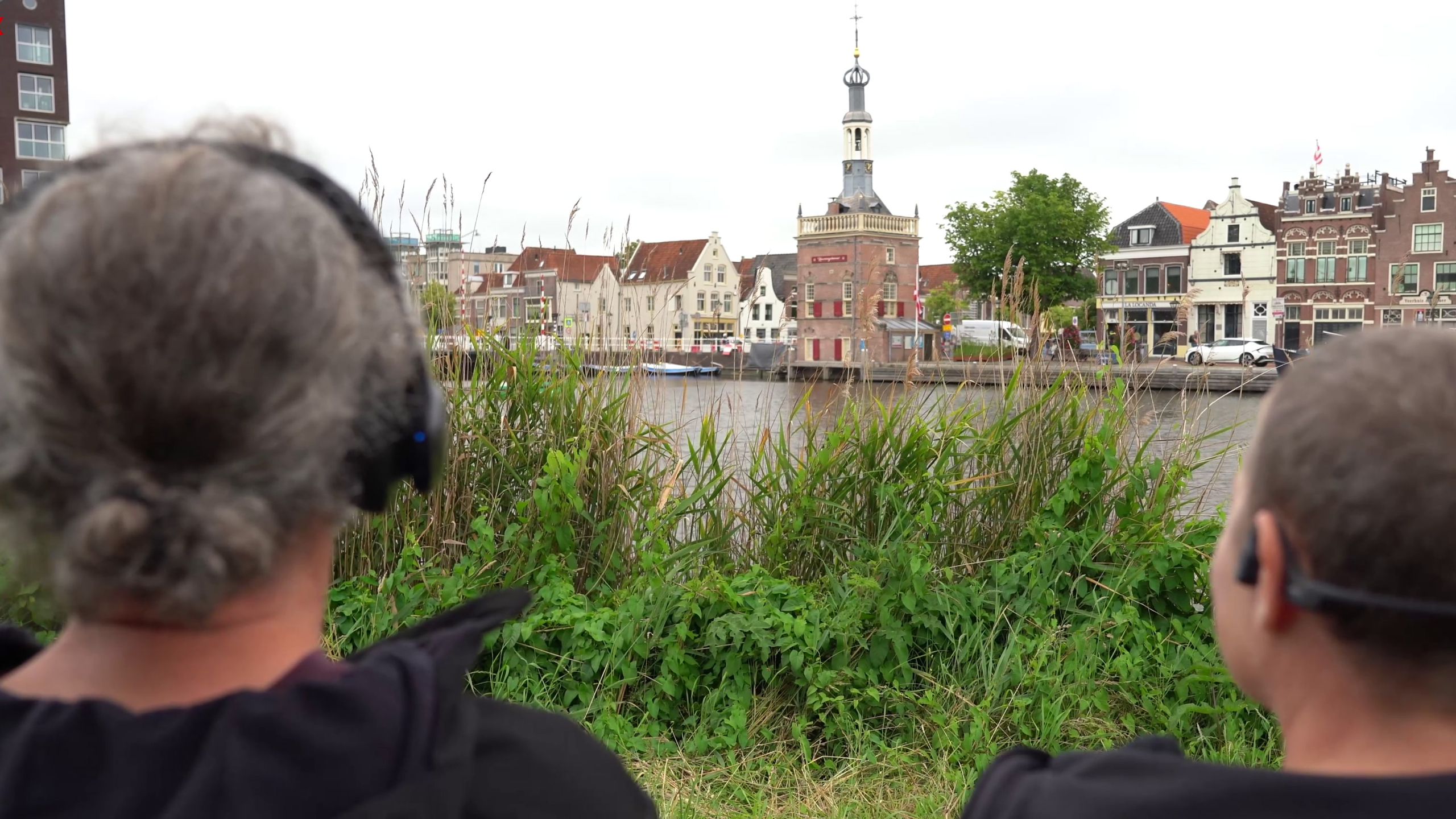 Twee personen met hoofdtelefoons zitten op de voorgrond, kijkend naar de historische gebouwen en de toren aan de overkant van het water in een Nederlandse stad, met riet en gras op de oever.