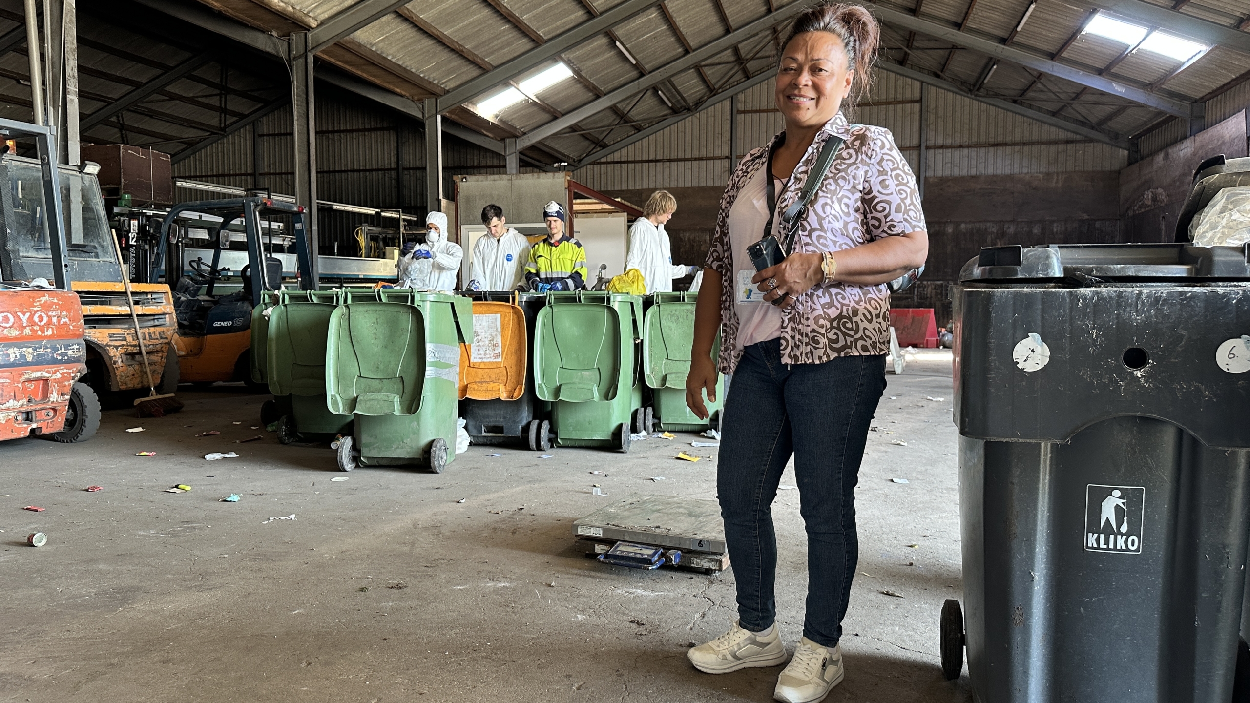 Een vrouw staat in een loods met verschillende groene afvalbakken op de achtergrond waar mensen in beschermende kleding mee bezig zijn.