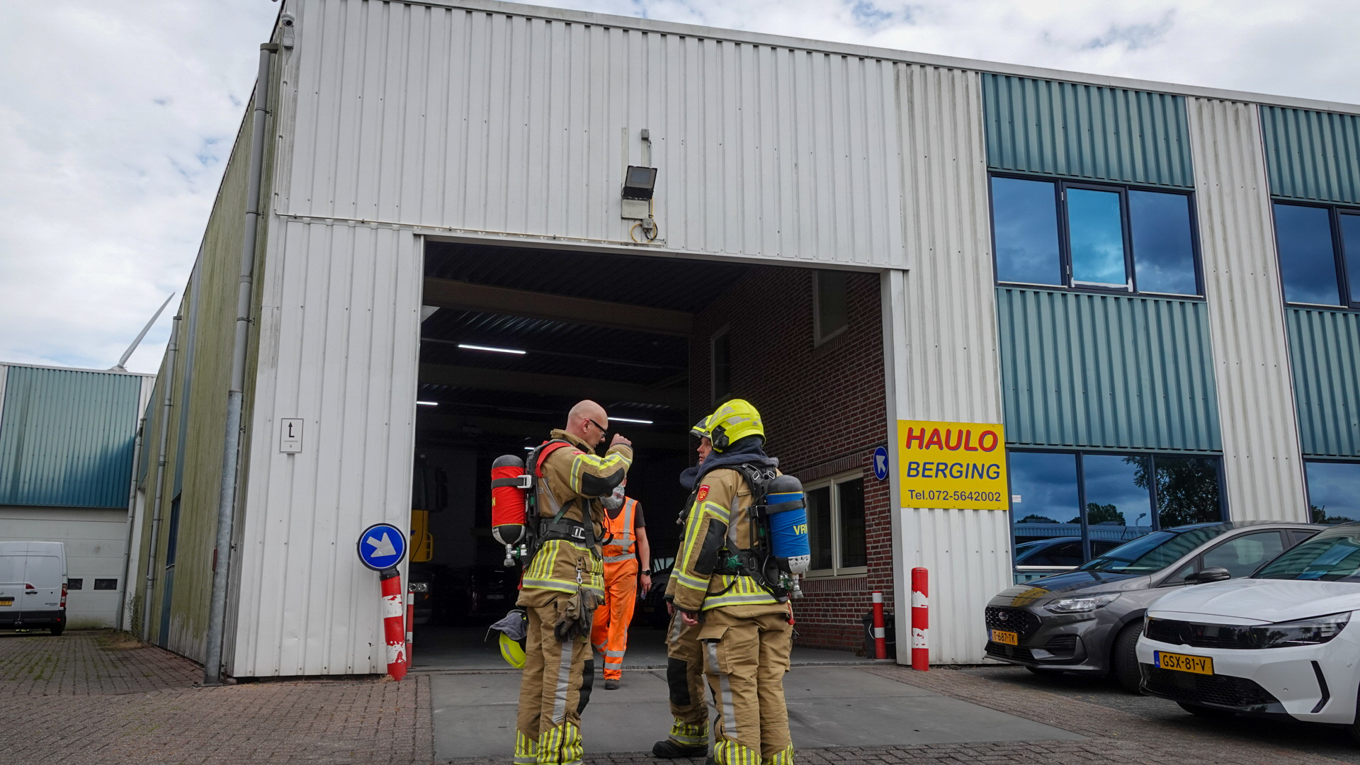 Brandweermannen in beschermende uitrusting staan voor een grote loods met op de achtergrond een persoon in een oranje veiligheidsoutfit. Rechts is een bord met 'HAULO BERGING' zichtbaar en geparkeerde auto's.