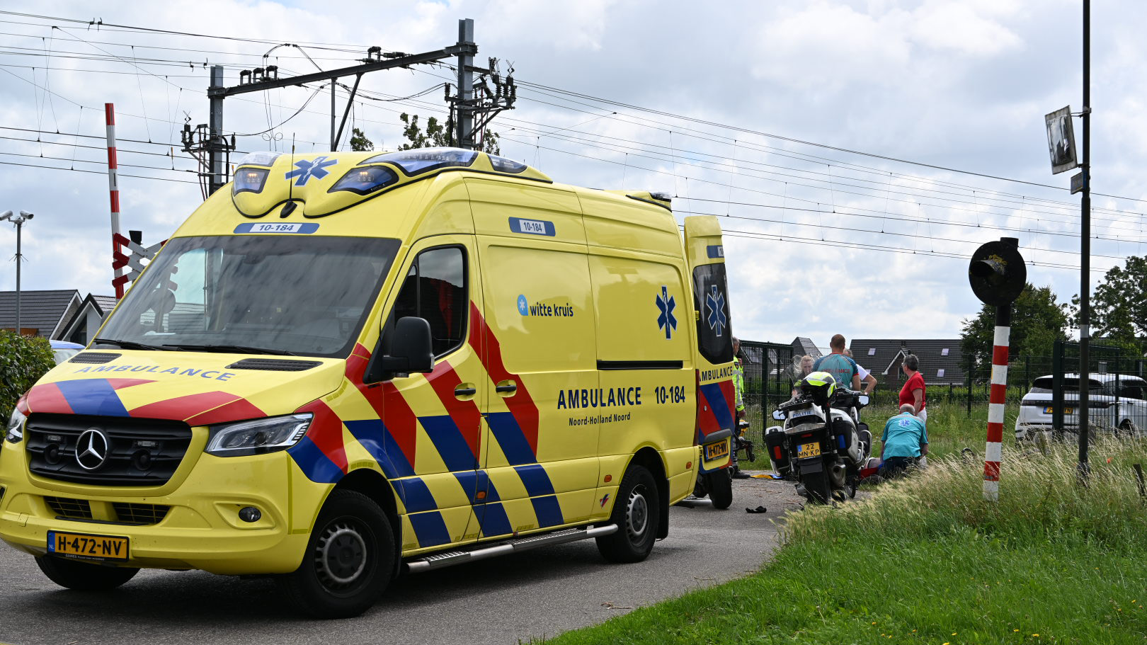 Gele ambulance staat op de weg bij een spoorwegovergang, waar hulpverleners bezig zijn met een persoon op de grond. Er is ook een motor van een medische dienst aanwezig.