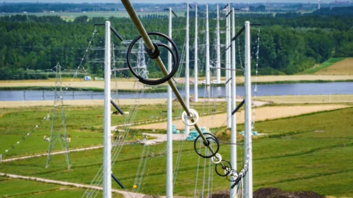 Hoogspanningslijnen die over een groen landschap en een rivier lopen, ondersteund door een rij metalen masten, met een bosrijke achtergrond op de horizon.
