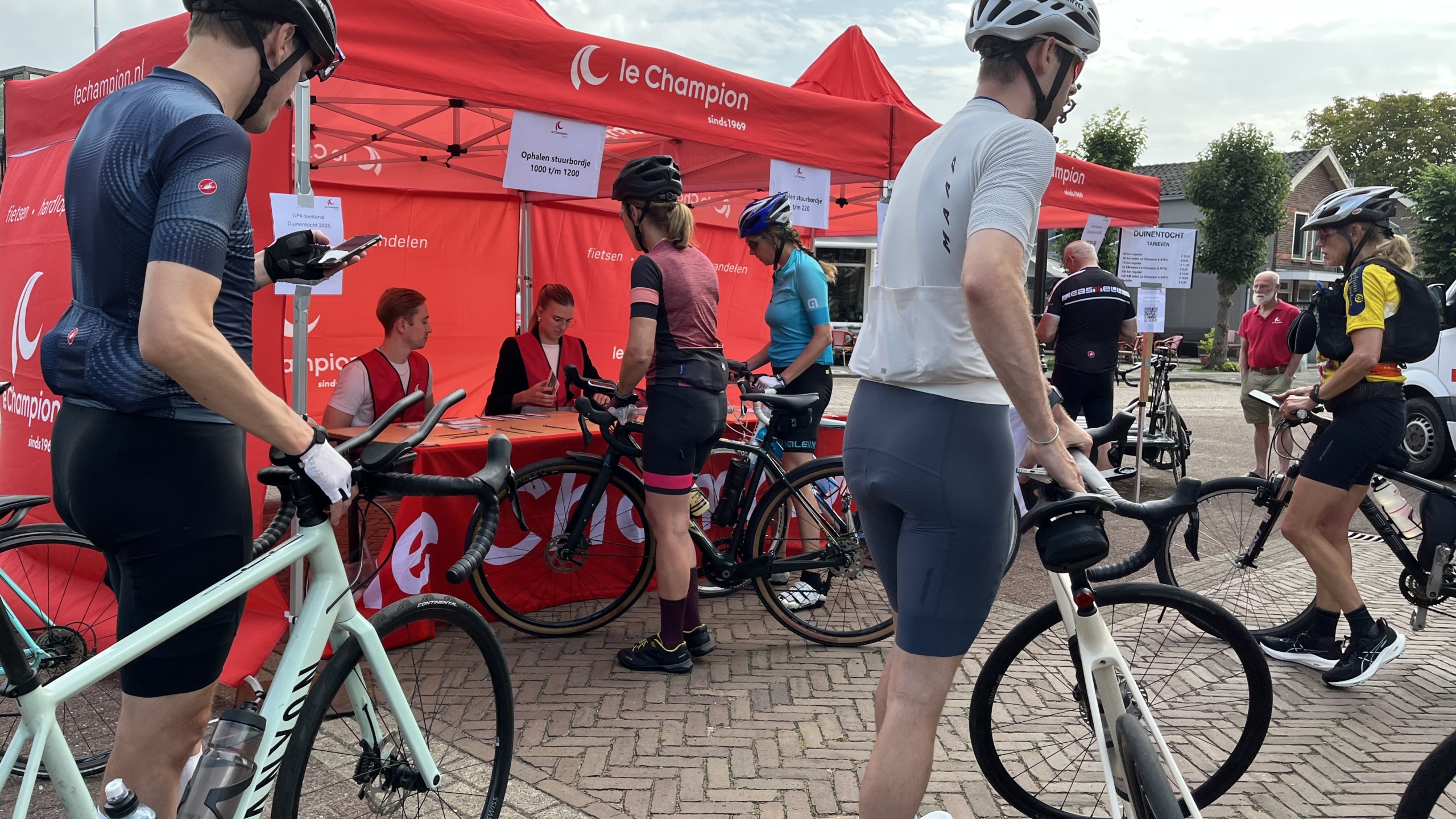 Fietsers verzamelen zich bij een rode tent van 'Le Champion', waar ze hun stuurbordjes ophalen. Twee medewerkers zitten aan een tafel onder de tent om de deelnemers te registreren. De fietsers dragen sportieve fietskleding en helmen. Op de achtergrond zijn huizen en bomen te zien.