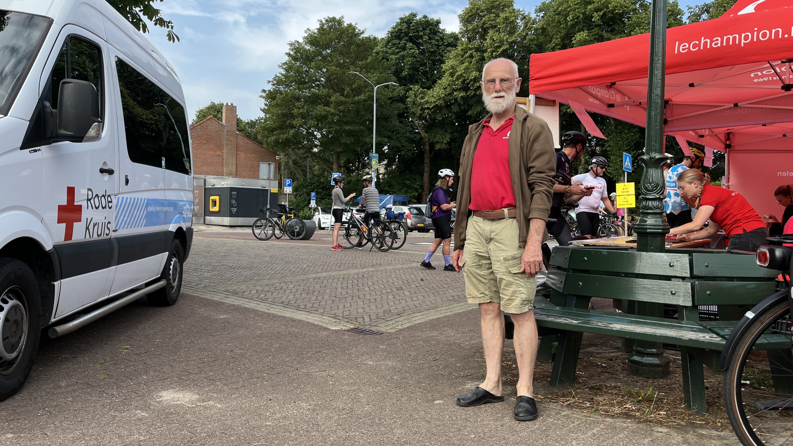 Oudere man in een rode poloshirt en korte broek staat voor een Rode Kruis busje naast een rood tentje, met fietsers en vrijwilligers op de achtergrond.